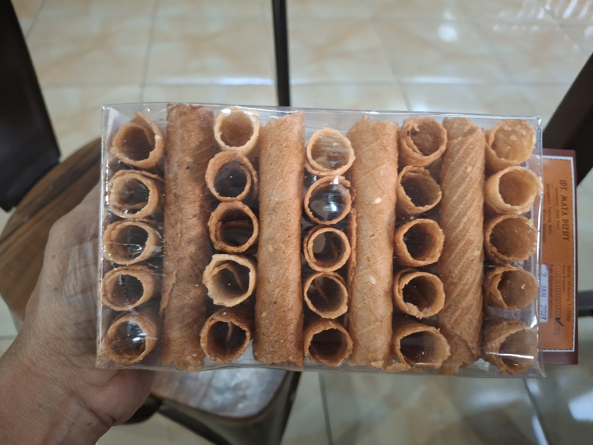 An overhead shot shows a transparent plastic container filled with rolled wafers. The container is placed on a light-colored tiled surface. The rolled wafers vary in their filling; some are hollow, while others contain a dark substance, likely chocolate. The container is oriented horizontally. To the right of the container, a portion of brown cardboard packaging is visible, displaying text including "IPT MAYA PIZIY" and a date that reads "2 JAN 2027". A person's left hand and arm, with visible skin texture and a ring on the index finger, is seen in the bottom left corner, holding the container. The background is blurred, showing hints of a tiled floor and furniture. The overall scene appears to be an indoor setting, likely a shop or home.
