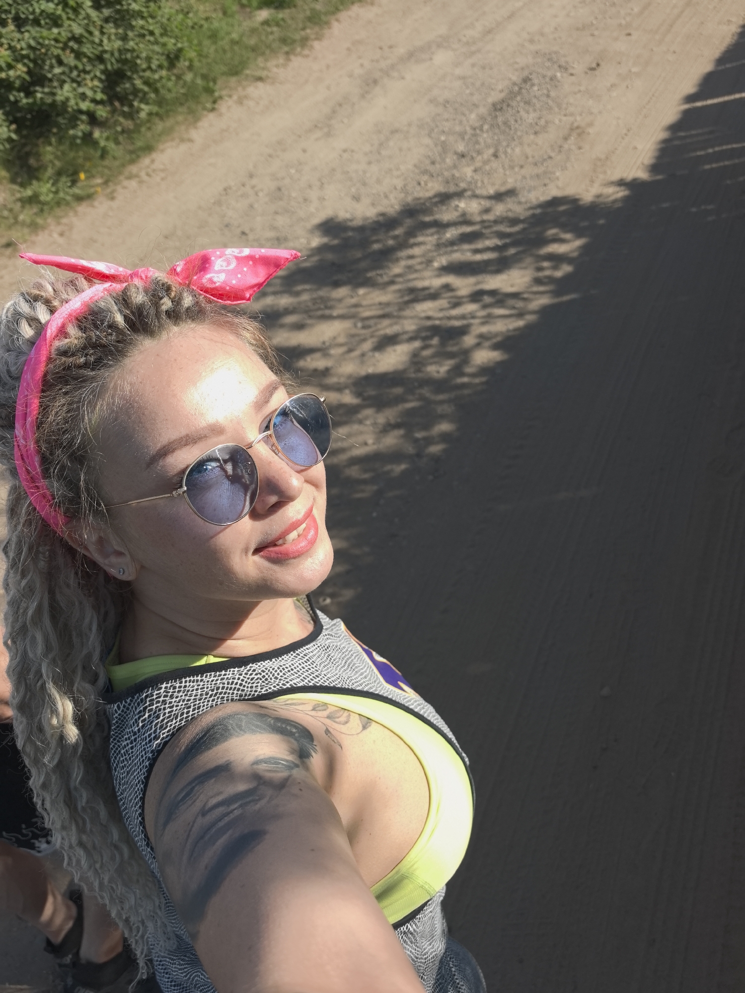 A blonde-haired woman with dreadlocks, a pink headband, and round sunglasses is taking a selfie. She's wearing a yellow and black patterned tank top and has a tattoo on her upper left arm. The background is a dirt road with vegetation on the left side and a shadow on the right. The image appears to be taken in direct sunlight. There's no indication this image was taken in Noviy, Russia.