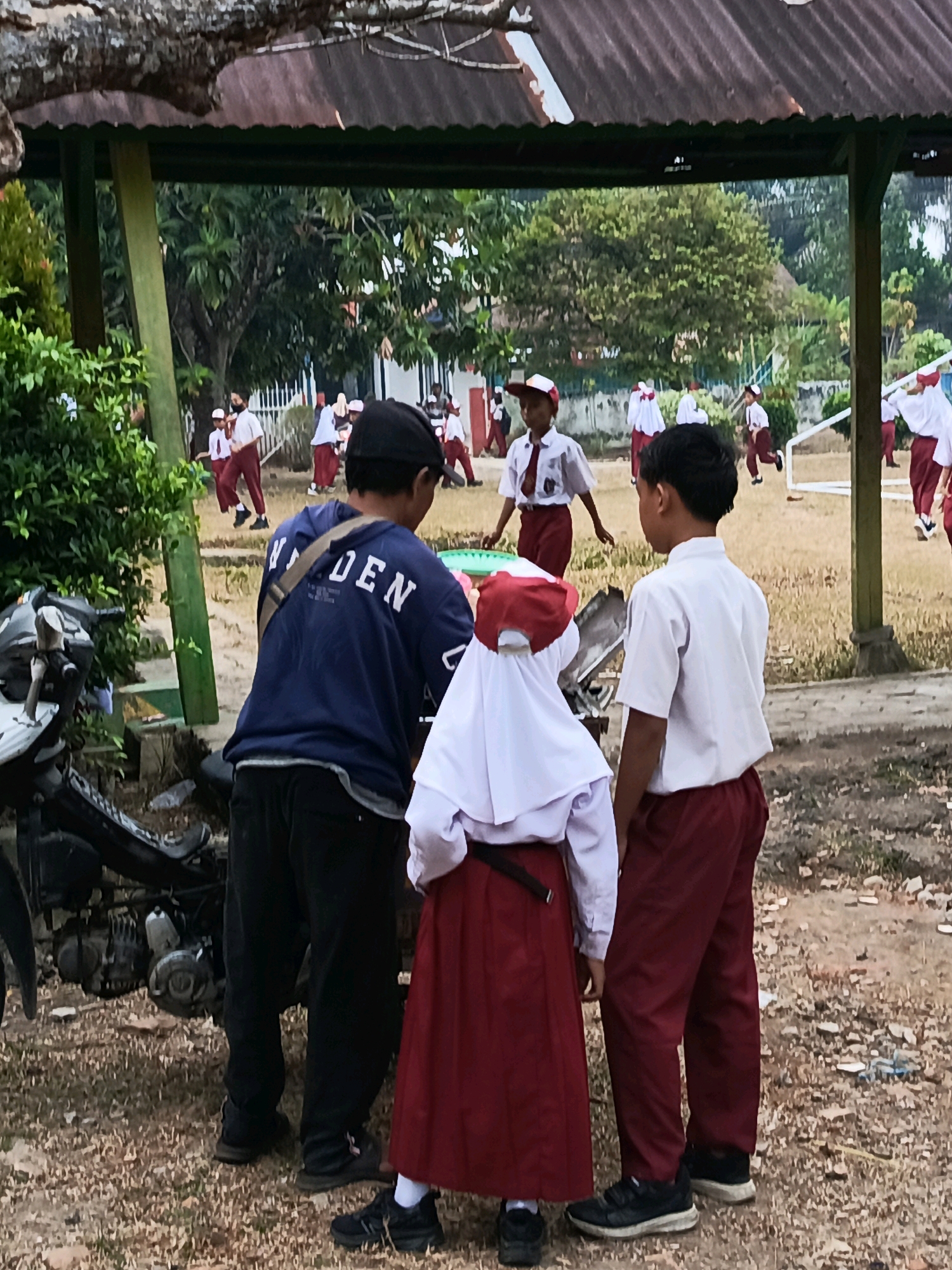 In the city of Megang Sakti II, Indonesia, an outdoor scene unfolds beneath a covered structure, possibly a school pavilion. A man wearing a dark blue hoodie with white lettering and black pants stands facing away from the camera, his back to a parked motorcycle. He has a strap from a bag slung across his back. To his right, a young boy in a white short-sleeved shirt and maroon pants, with black hair, looks away from the viewer. In front of the man and to the right of the boy, a young girl wearing a white hijab and a maroon skirt stands, also facing away. In the background, several children in similar white shirts and maroon pants, some wearing white headscarves, are dispersed across a dusty field. Some children appear to be running or engaged in activities. The environment is characterized by dry ground, scattered debris, and lush green foliage from trees and bushes.