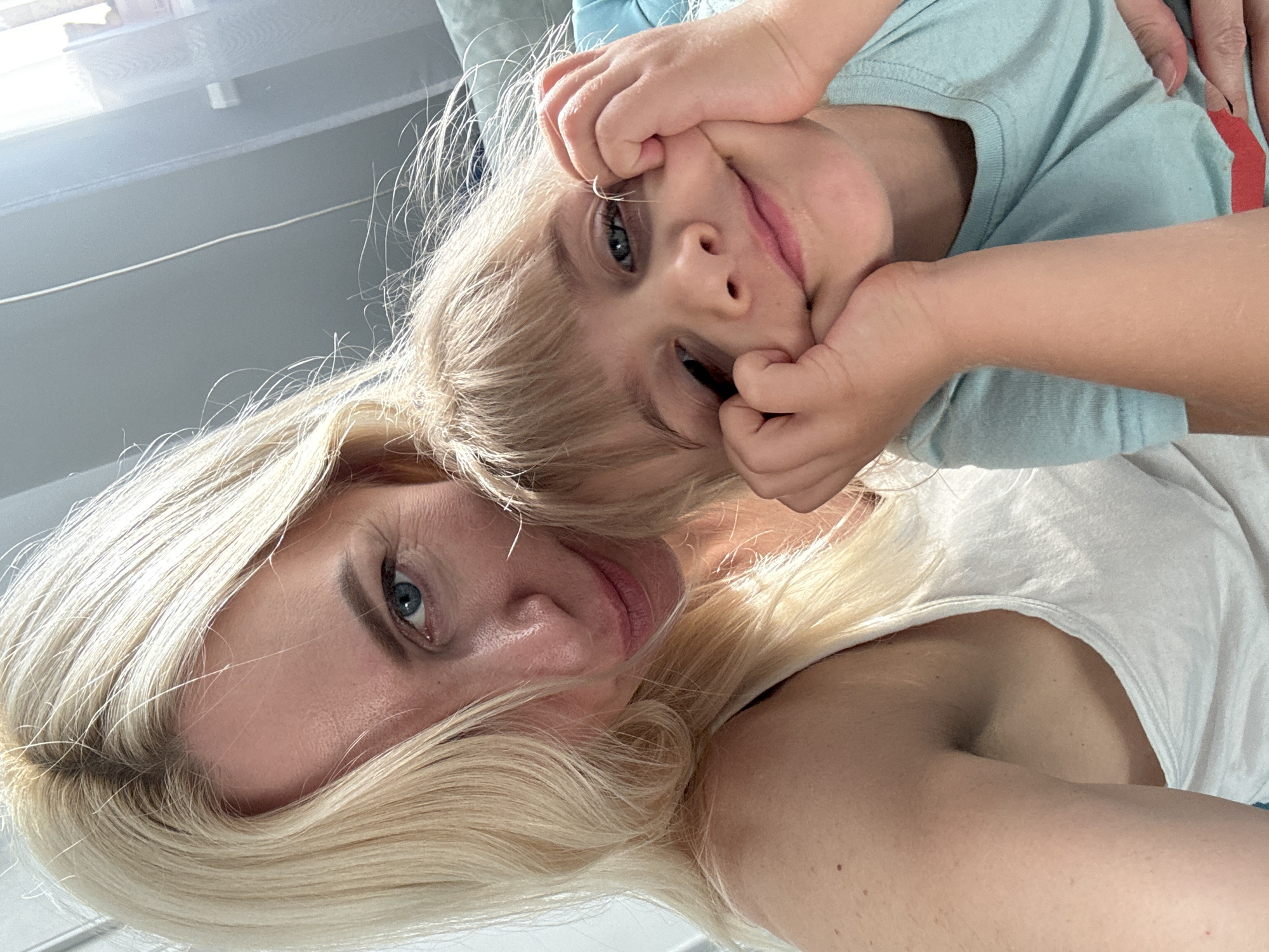 A close-up, selfie-style photograph captures two individuals, a woman and a young child, in what appears to be an indoor setting. The woman, positioned on the left, has blonde hair and blue eyes, and is wearing a white tank top. She is looking towards the camera. The child, on the right, also has blonde hair and is making a playful face, with hands propping up their cheeks. The child is wearing a light blue t-shirt. The background is slightly blurred, showing what could be a window with sheer curtains on the right and a neutral-colored wall or furniture on the left. The lighting suggests it is daytime, possibly with natural light filtering in. There is no visible indication of the city of Unknown, Russia.