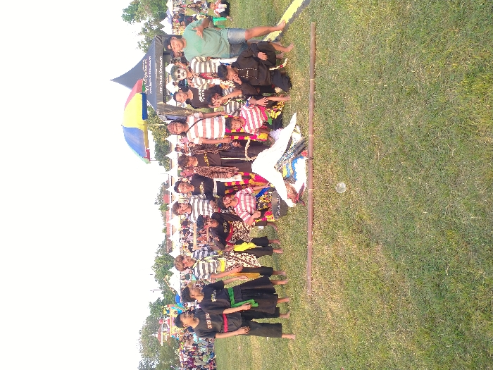 The image depicts an outdoor gathering of approximately 18-20 individuals, predominantly young males, on a grassy field in Glonggong, Indonesia. In the immediate foreground, one individual lies prone, facing away from the camera, dressed in a white top and a patterned lower garment. Two parallel bamboo poles are positioned on the grass in front of this prone individual. Surrounding them, other individuals are standing or kneeling. Attire includes striped long-sleeved shirts, dark pants, patterned sarongs, and some individuals displaying white theatrical face paint. One person on the far right wears a light green t-shirt and blue shorts. The background features multiple white peaked tents, a large black conical tent with discernible text, and a multi-colored umbrella. Distant crowds and trees are visible along the horizon under a bright sky, indicating a public event.