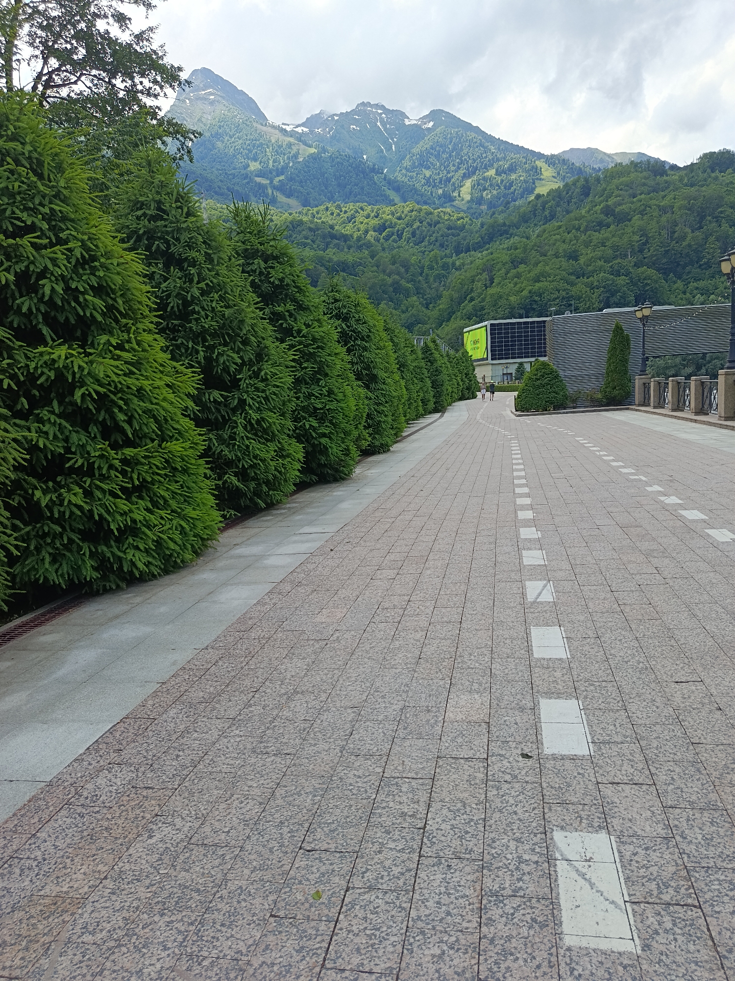The image depicts a pathway in Estosadok, Russia, leading towards a modern building complex nestled at the base of lush, forested mountains. A row of manicured coniferous trees lines the left side of the pathway, their dense foliage creating a vibrant green contrast against the muted tones of the pavement and sky. The pathway itself is paved with large, speckled granite tiles, with a lighter colored, textured sidewalk running parallel to the trees. White rectangular markings are painted on the granite path, suggesting a designated lane or walking area. In the background, the mountains are covered in dense greenery, with patches of snow visible on the higher peaks, indicative of a summer or late spring scene. The sky is overcast, with a mix of gray clouds and lighter patches. A modern building with large windows and a distinctive green sign is visible to the right, partially obscured by the trees and mountains. Two individuals are walking away from the camera on the pathway in the distance. The overall impression is one of a well-maintained urban or resort area integrated with a natural mountain landscape.