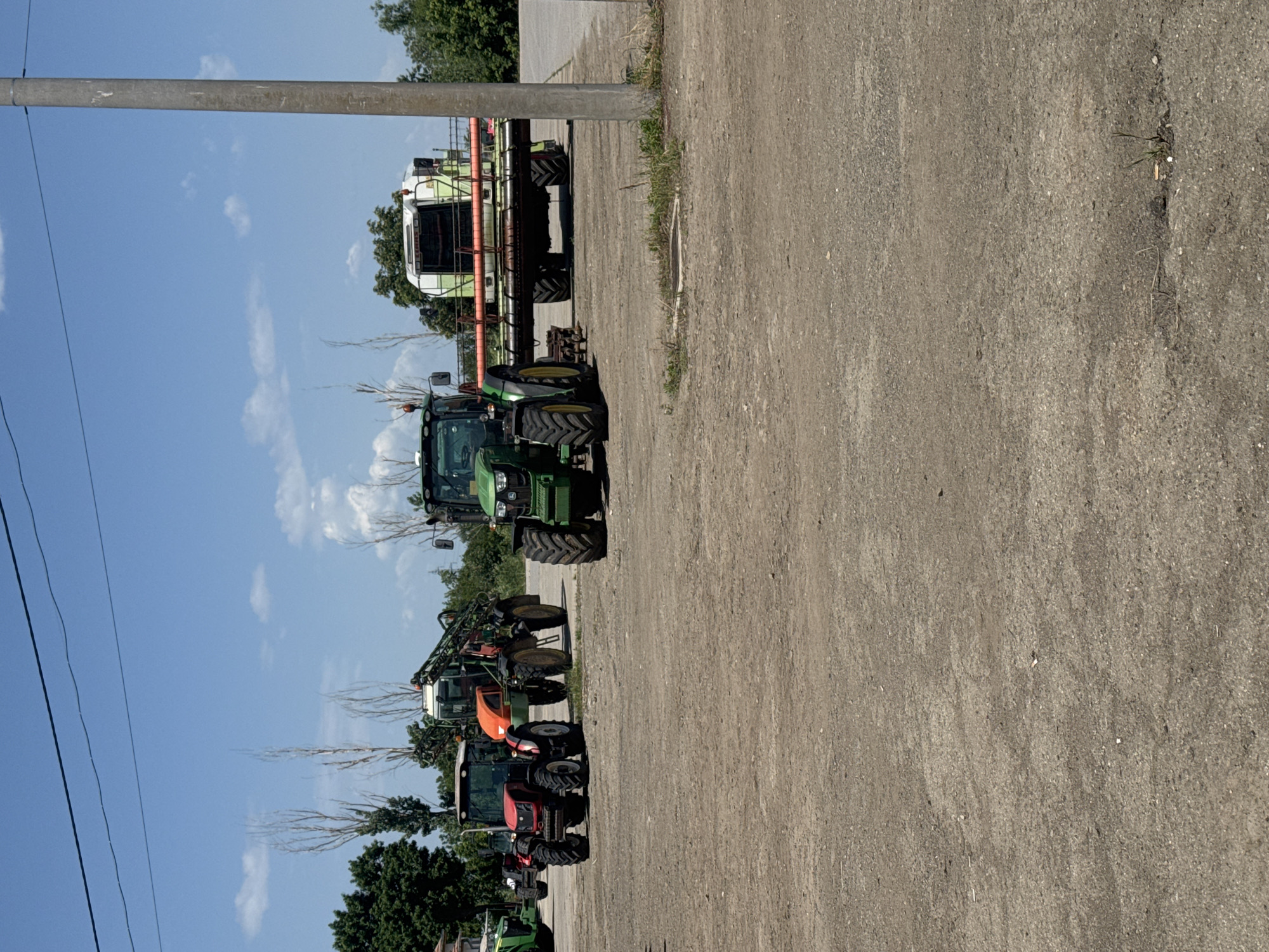 The image shows a row of parked agricultural vehicles on a paved surface, likely a parking lot or storage area. There are multiple tractors, some with attached implements such as sprayers, along with a combine harvester at the end of the row. The vehicles are of different colors, including green, red, and orange. A utility pole is visible near the front of the image. The sky is blue with some clouds. Based on the prompt, this scene is located in Sarnevo, Bulgaria.