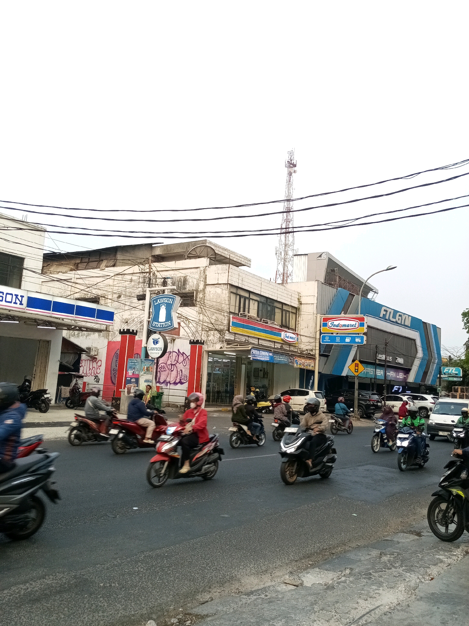 This is an eye-level, medium shot of a busy street scene in Bekasi, Indonesia, on a cloudy day. The foreground shows a paved road with various motorcycles and their riders in motion, some blurred due to movement. To the left, a partial view of a black motorcycle is visible. In the mid-ground, numerous motorcycles are traveling on the road, many with riders wearing helmets and some with passengers. Buildings line the street. On the left side, a white building with blue trim has a sign that reads "LAWSON STATION". Adjacent to it, another building features red and white striped pillars with graffiti. Further to the right, a larger building with a blue facade displays a prominent sign for "FTL GYM". Other signs visible include "Indomaret" and what appears to be a traffic signal. A cellular tower is visible in the upper right portion of the image. The overall impression is one of bustling urban activity.