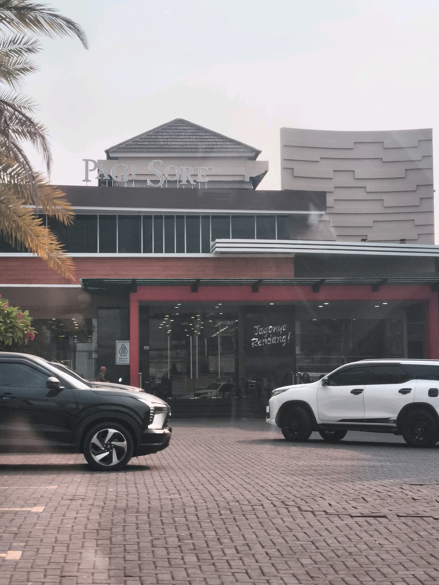 A frontal, slightly low-angle shot captures the facade of a restaurant, "PAGI SORE," in Palembang, Indonesia, on a bright, hazy day. The building features a distinctive tiered, curved facade on the right and a darker, horizontal section with windows above a vibrant red entrance. A prominent sign with silver, raised lettering reads "PAGI SORE." To the left of the entrance, a white sign with a geometric black symbol is visible. A large palm frond arches over the top left of the frame. In the foreground, a dark SUV is parked on a brick-paved surface, its front left wheel turned, and a white SUV is parked on the right side of the frame. The interior of the restaurant is visible through large glass windows, showing tables and lighting. The street is paved with interlocking brick pavers, with faint yellow lines marking parking spots. The sky is a uniform, pale grey, suggesting overcast conditions or strong haze.