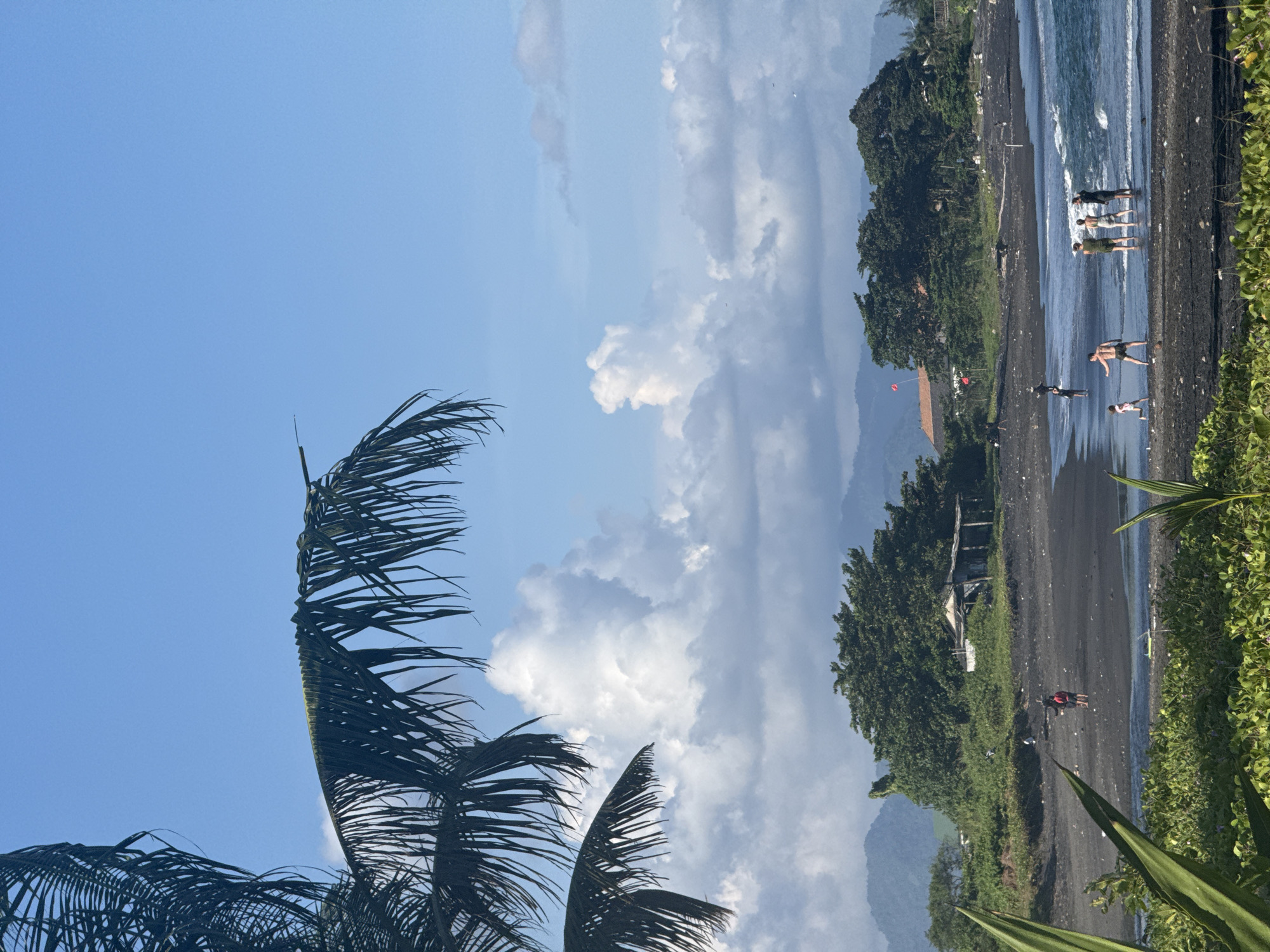 A vibrant beach scene in Medahan, Indonesia is captured under a partly cloudy sky. The black sand beach stretches along the coastline, where several people are wading in the surf or walking along the shore. Lush green vegetation lines the edge of the beach. Palm fronds frame the scene on one side, adding a tropical element. In the background, a mountain range is visible.