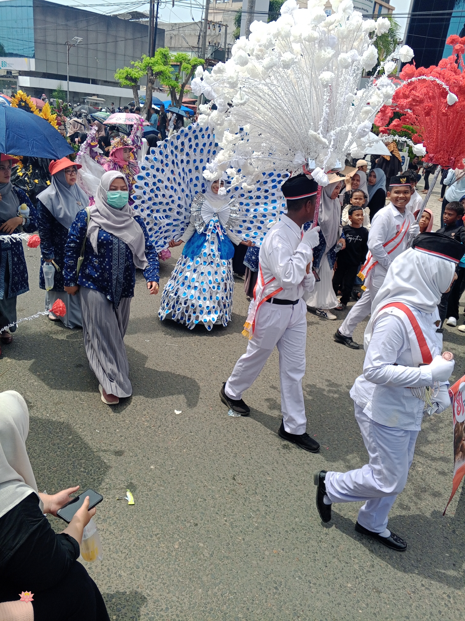 This is a medium shot, eye-level, outdoor photograph taken during a parade in Lhokseumawe, Indonesia. The central focus is on a group of people dressed in elaborate costumes, participating in a parade on a paved road.
In the foreground, a person in a white uniform with a red sash walks towards the right, holding a silver object. To their left, another individual in a similar white uniform also walks right, holding a wooden stick. Behind them, a woman in a grey hijab and a blue patterned long-sleeved shirt walks to the left, wearing a green face mask and a long grey pleated skirt. She is holding a transparent plastic bag with yellow objects inside and a smartphone. To her left, another woman in a grey hijab and blue patterned clothing is visible.
Dominating the center and extending to the right is a large, ornate costume resembling a peacock, with a fan-like tail made of white paper-like material, adorned with blue circular patterns. A person in a blue and white costume, possibly part of the peacock outfit, is partially visible beneath it. To the right of the peacock costume, a large, red, elaborate decoration is visible, also appearing to be part of a costume or float.
In the background, numerous other parade participants and spectators are visible, some holding umbrellas to shield themselves from the sun. Buildings, including one with the "BSI" logo, line the street. Trees and utility poles are also present in the background, indicating an urban setting. The ground is asphalt, with scattered debris and shadows cast by the people and structures. The overall atmosphere suggests a festive community event.