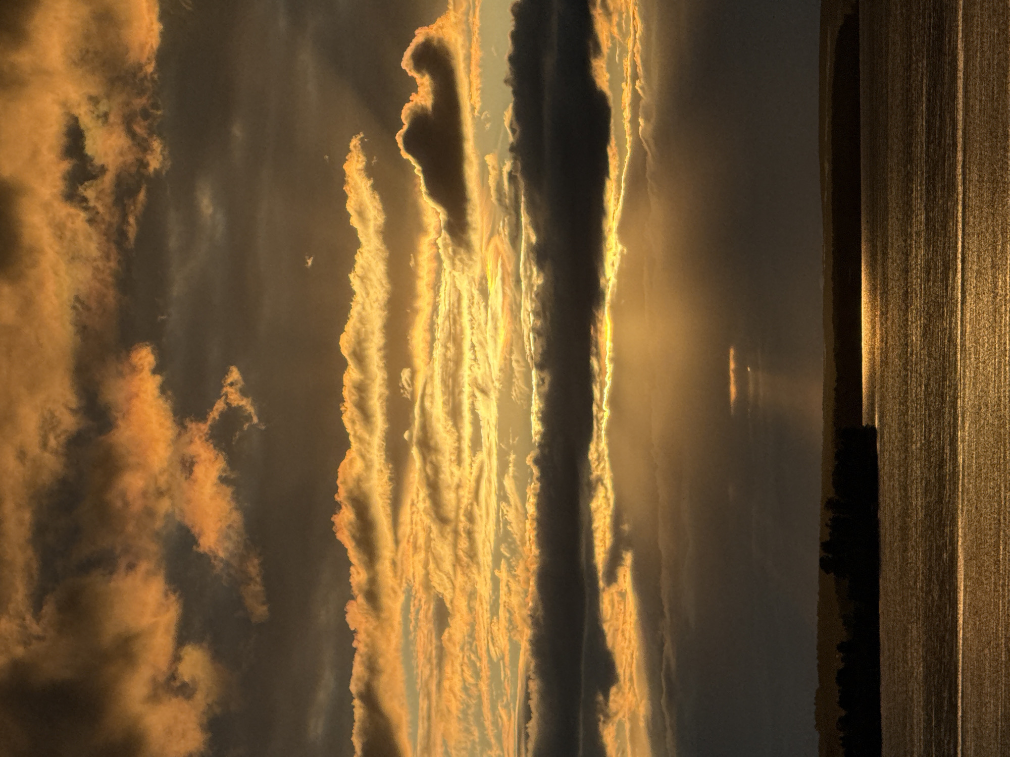 This media file depicts a dramatic sunset over a body of water, likely a lake or river, with a dark tree line visible in the distance. The sky is filled with a turbulent display of clouds illuminated by the setting sun. The clouds exhibit a range of colors, from deep grays and blacks to vibrant oranges and yellows, with streaks of light piercing through them, creating a sense of awe and grandeur. The sunlight reflects off the water's surface, creating a shimmering, golden path that stretches towards the horizon. The overall atmosphere is one of natural beauty and powerful, elemental forces. The image's orientation is vertical, suggesting it was captured in portrait mode. The scene evokes a feeling of solitude and reflection, as one might experience while witnessing such a striking natural phenomenon. The time of day is clearly sunset, given the intense coloration of the sky and the low angle of the sun. The weather appears to be a mix of clear skies and cloud cover, contributing to the dramatic visual effect. No text is visible in the image. The location context of Votkinsk, Russia, suggests a northern European landscape, though specific geographical features are not distinctly identifiable beyond the water and trees.
