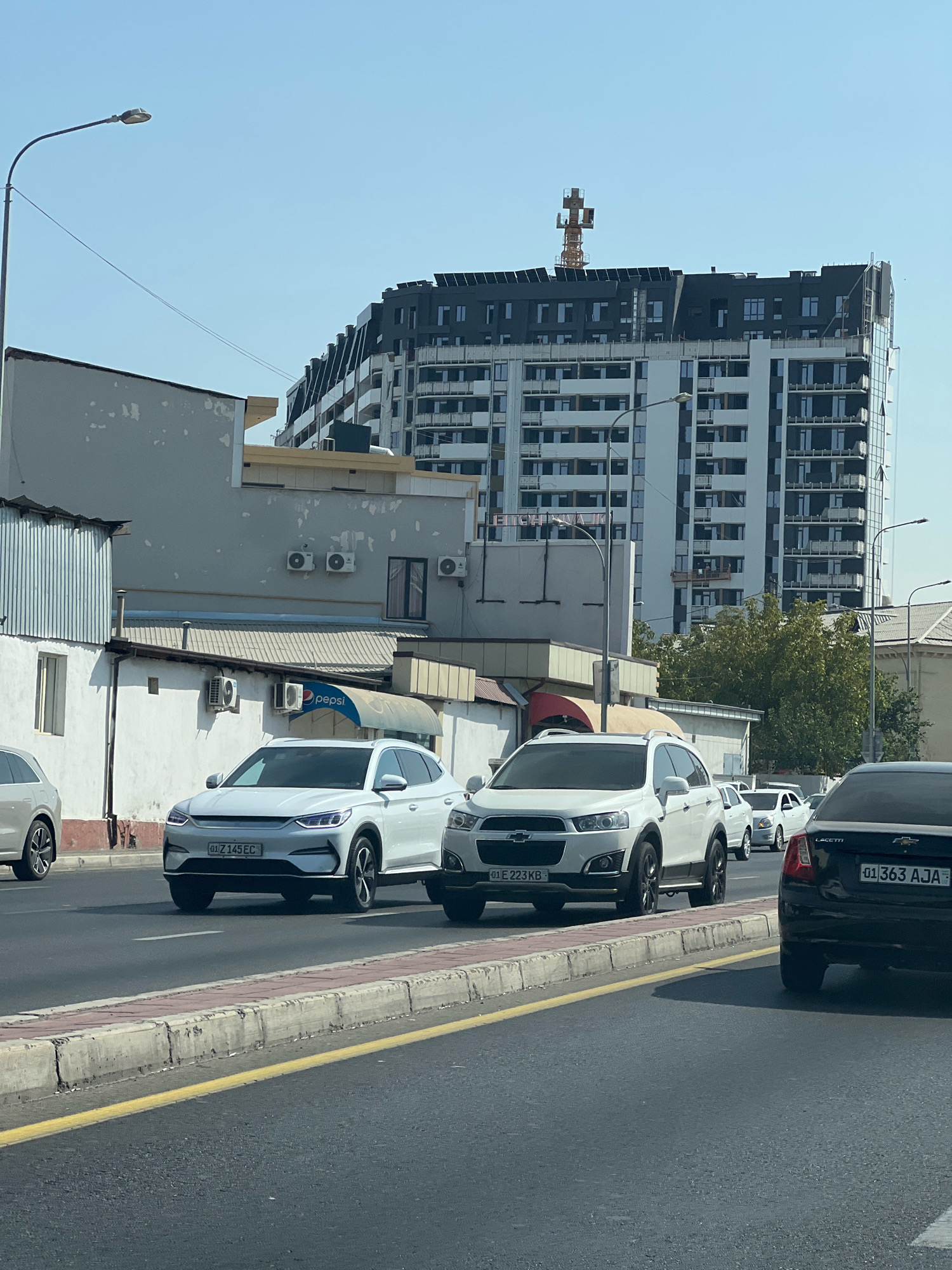 The image shows a street scene in Tashkent, Uzbekistan, on a clear day. In the foreground, a multi-lane asphalt road is visible, with a concrete and brick curb separating it from a sidewalk. Several cars are driving on the road, including a white BYD electric vehicle with license plate "01 Z 145 EC" in the center lane. To its right, a white Chevrolet Captiva with license plate "01 E 223 KB" is in the right lane. Further down the road, a black Chevrolet Lacetti with license plate "01 363 AJA" is visible.
The middle ground features a collection of buildings. On the left, a single-story building with a light gray, weathered facade and a corrugated metal roof section is present. Next to it, another light gray building shows signs of wear, with visible air conditioning units mounted on its exterior. A large Pepsi logo is partially visible on an awning. In the background, a tall, modern apartment building with a distinctive dark-colored, stepped facade dominates the skyline. A construction crane is visible on top of this building. Trees and other smaller buildings are also interspersed within the urban landscape. The sky is a clear, light blue.