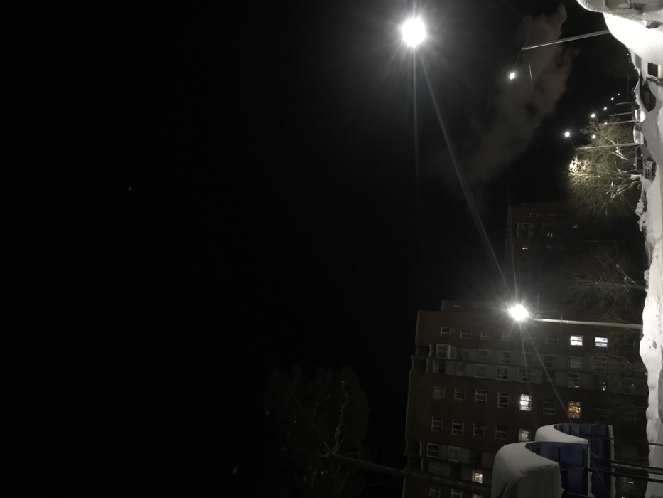At night, a tall apartment building with lit windows stands next to a dark building in the background. Street lights illuminate the scene, their bright beams cutting through the darkness and revealing snow-covered surfaces. Bare trees can be seen near the buildings. Some clouds are visible above the buildings.