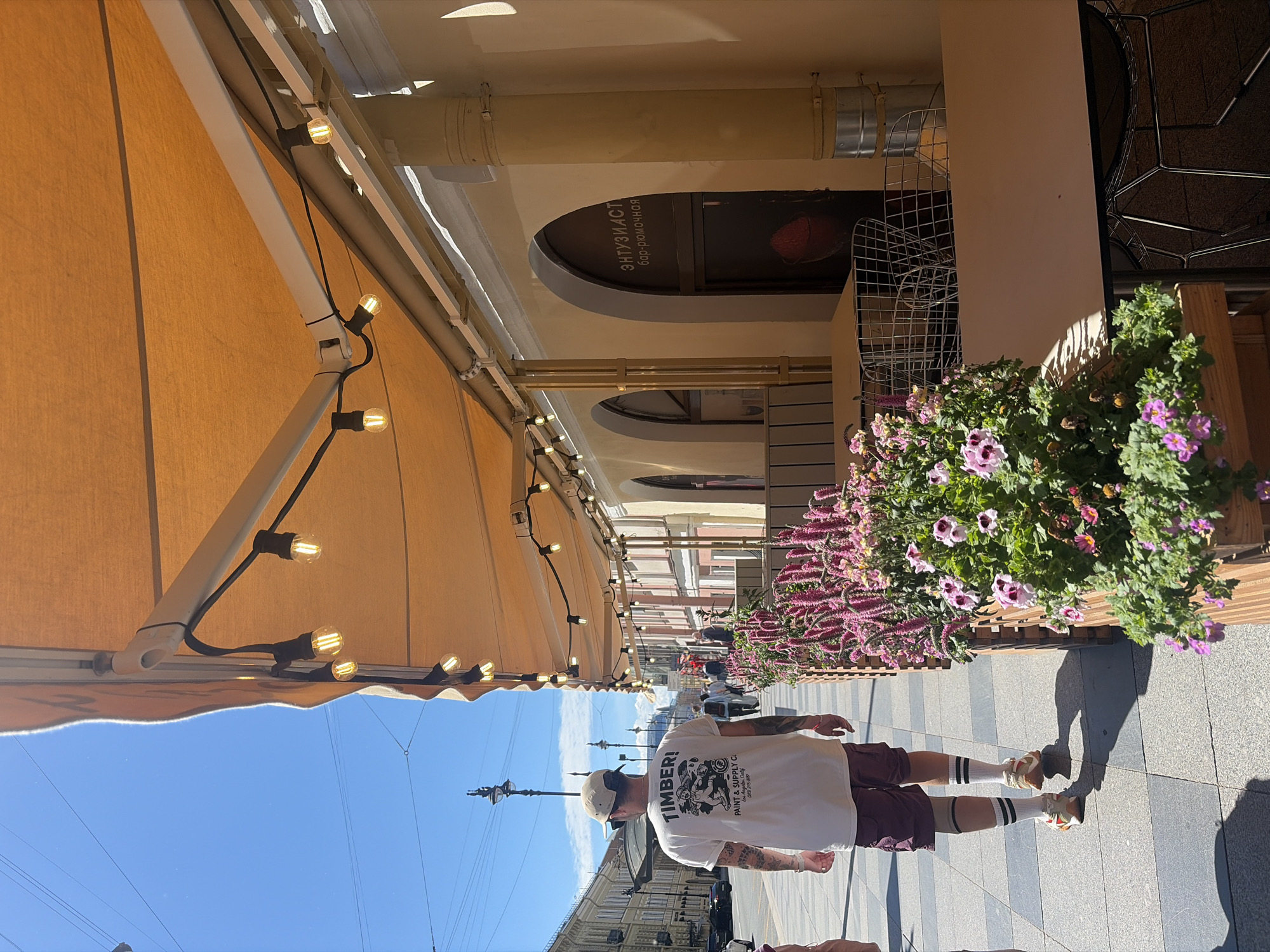 This is a vertical, eye-level, medium shot of a man walking down a sidewalk in Saint Petersburg, Russia. The man is wearing a white t-shirt with graphics, burgundy shorts, white socks with black stripes, and sneakers. He has tattoos on his arms and is wearing a baseball cap. The sidewalk is paved with gray and black tiles, and there is a wooden planter box filled with pink and purple flowers next to the sidewalk. In the background, there are buildings with arches, tables and chairs set up for outdoor dining, and string lights with Edison bulbs hanging from an orange awning. The sky is blue with some clouds.