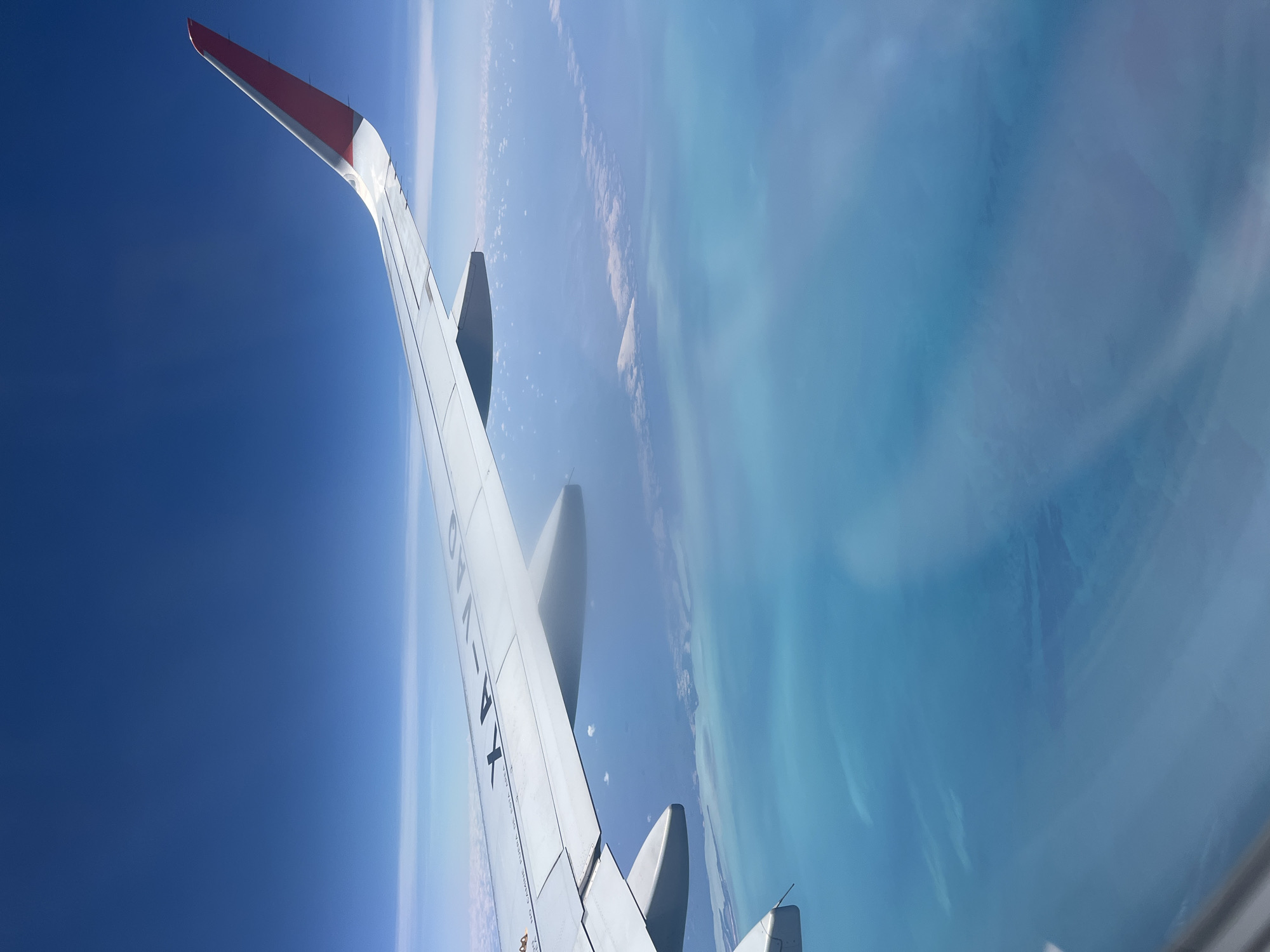 An aerial view captures the wing of an airplane against a backdrop of clear blue sky and vibrant tropical waters. The white aircraft wing, featuring a red and white wingtip, extends diagonally from the upper left. Below the wing, the ocean presents stunning hues of turquoise and light blue, indicative of shallow, clear waters over reefs or sandy bottoms. Irregular landmasses or islands are visible along a coastline, contrasting with the lighter water. The sky above is a deep, cloudless blue, suggesting bright, clear daytime conditions. Two engine nacelles are partially visible beneath the wing. The image appears to be taken from a passenger window, with subtle reflections or distortions visible on the glass.
On the upper surface of the wing, the aircraft registration "XA-VAQ" is clearly printed in black. Additionally, part of a warning message, "NO CAMINE FUERA DE...", is visible below the registration. The scene conveys the experience of flying over a beautiful coastal region. Given the location context of Ukum, Mexico, this view likely depicts the Caribbean coast or the Yucatán Peninsula.