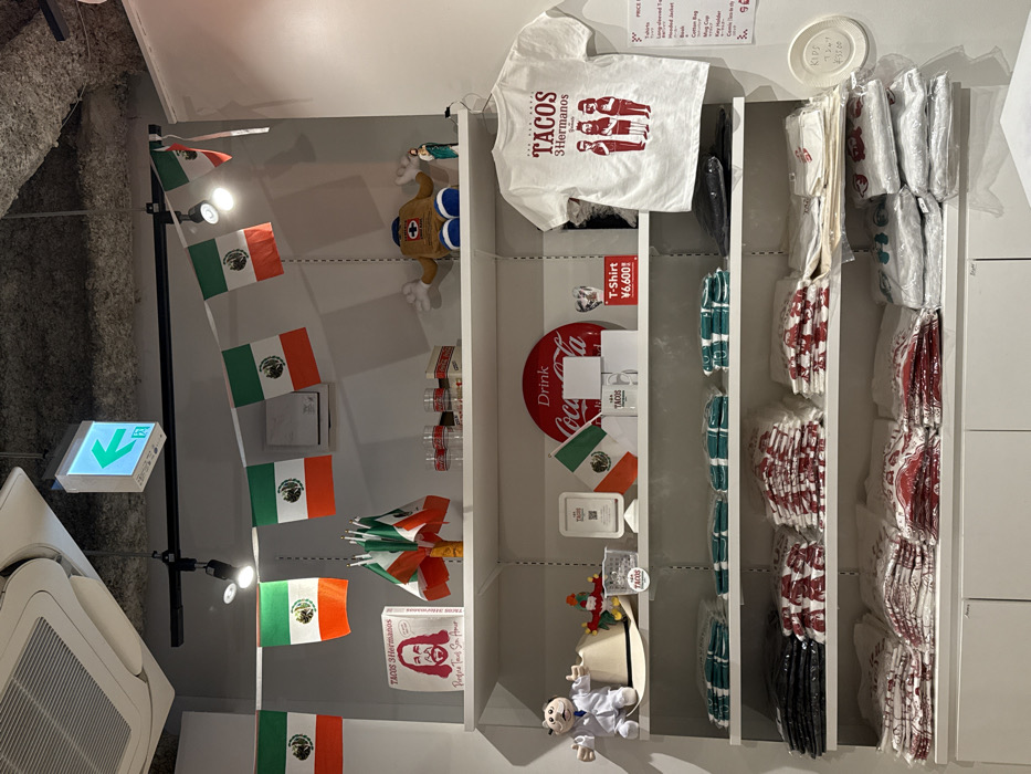 This image depicts a brightly lit indoor retail display, confirmed to be located within a commercial establishment in Shibuya, Japan. The ceiling features exposed concrete, track lighting fixtures, and an illuminated green emergency exit sign with a left-pointing arrow and Japanese text.
A white shelving unit spans the width of the frame, organized with various merchandise. Mexican flag bunting hangs above the top shelf. On the top shelf, from left to right, are a square white box with "Tacos 3 Hermanos" branding and a stylized image of Jesus, a cluster of small Mexican stick flags, multiple clear glasses with a red Coca-Cola logo, and various decorative items including a brown plush figure, and a small green figurine.
The second shelf from the top displays a white plush figure, a light brown cowboy hat, a small Mexican flag, a white stand with a QR code, a round red "Drink Coca-Cola" sign, white mugs, and a hanging white t-shirt with "Tacos 3 Hermanos" branding and three stylized figures, priced at ¥6,600 (Japanese Yen).
The third and fourth shelves below are stacked neatly with folded t-shirts, many individually packaged in clear plastic. These shirts appear in various colors including green, red, white, and black, displaying different designs. A small circular white sign on the right, affixed to the shelf, shows additional pricing information in Japanese, including "T-SHIRTS ¥3,500". The bottom of the image shows the top of white cabinetry or storage units beneath the shelves. No individuals are visible in the scene.