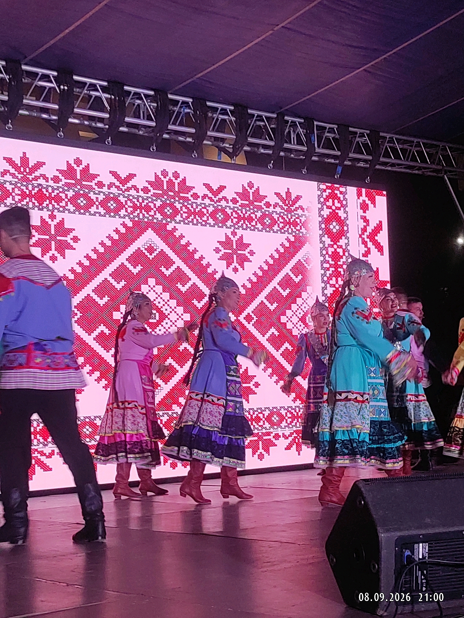 A stage performance is captured in Makhachkala, Russia, on September 8, 2026, at 21:00. The scene features several individuals dressed in traditional attire. A performer in the foreground on the left, partially visible, wears a blue and red patterned top and black pants with black boots. To their right, a performer in a pink and multi-colored dress with ornate embroidery and brown boots is in motion. Further to the right, a performer in a turquoise dress with similar intricate patterns and brown boots also appears to be dancing. Behind them, other performers in traditional costumes can be seen, suggesting a group performance. The backdrop is a large LED screen displaying a vibrant, repeating red and white pixelated pattern, reminiscent of folk embroidery. Above the screen, stage lighting and truss structures are visible. The stage floor is reflective and appears to be wooden. A large black speaker is positioned in the lower right corner of the frame.