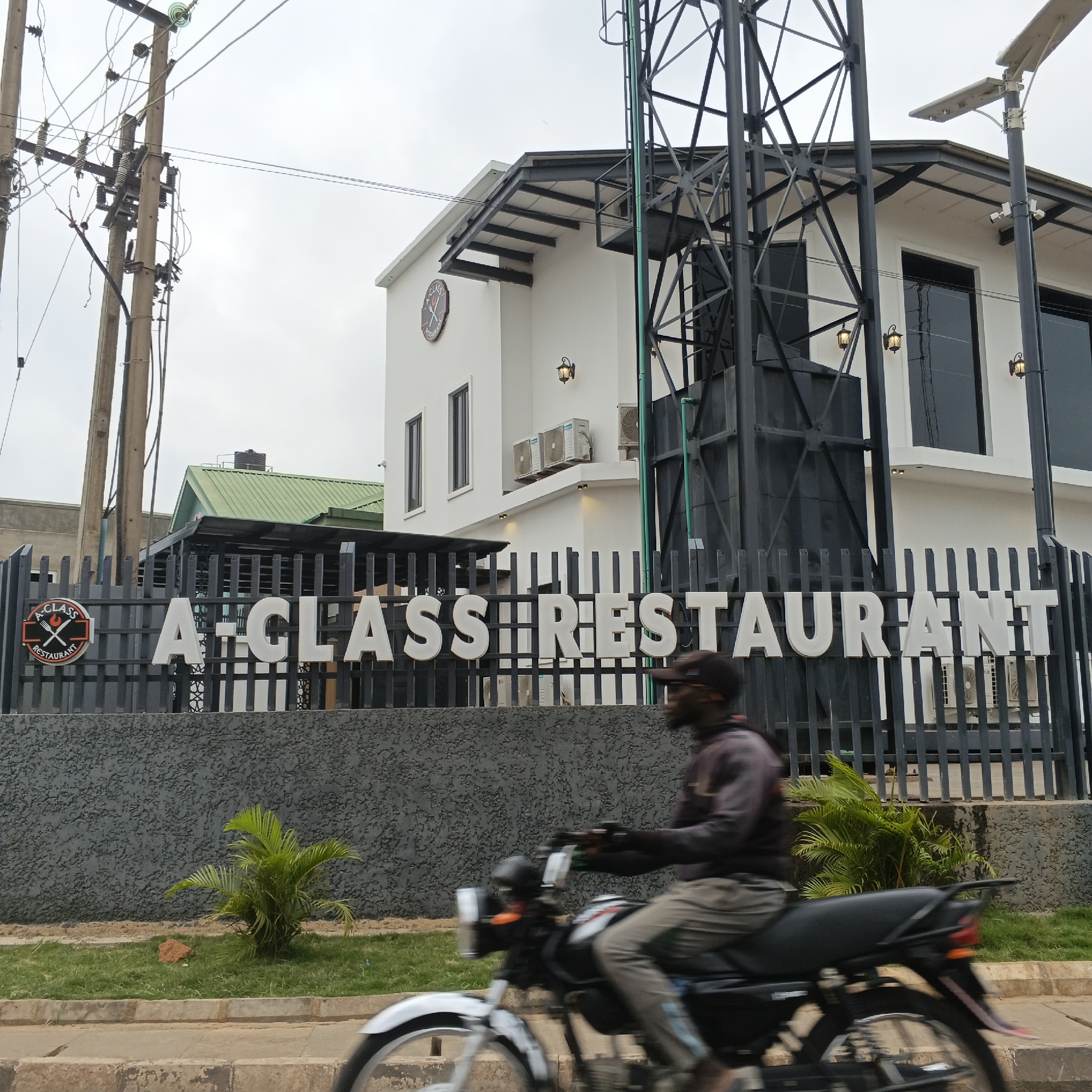 The image shows the exterior of "A-CLASS RESTAURANT" in Kabala, Nigeria. The restaurant building is white with dark trim and large windows. A black metal fence in front of the building displays the restaurant's name in large white letters and a circular logo with cutlery. A man on a motorcycle is blurred as he rides past in the foreground. Overhead power lines and poles are visible to the left, and a green-roofed building is partially visible behind the restaurant. The sky is overcast.