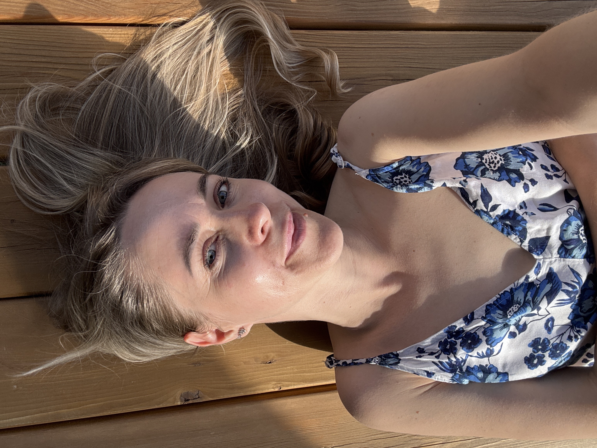 A woman is lying on a wooden surface. She has blonde hair and is wearing a white top with blue floral patterns. The surface appears to be a wooden deck or floor. The lighting suggests it is outdoors, possibly during the day. There is a distinct shadow cast across her hair and the wooden surface. There's no indication to ascertain the location to be in the city of Chaikovsky, Russia.