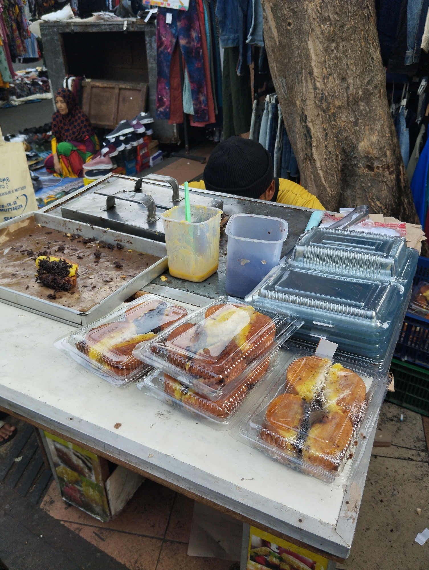 An outdoor food stall displays several loaves of bread, likely sweet bread or cake, packaged in clear plastic containers. The bread appears to have a golden-brown crust and some may contain chocolate or other fillings. Some loaves are whole, while others are sliced, revealing the interior. In the background, a person wearing a black beanie and a yellow shirt is partially visible, possibly the vendor, attending to the food. To the left, a woman wearing a headscarf and a colorful skirt is seated on the ground, possibly a customer or another vendor. The stall is set up on a street with a large tree trunk present, suggesting an urban environment in Surabaya, Indonesia. Various items of clothing are visible hanging in the background, indicating a market setting. The ground surface is rough, possibly pavement or dirt.