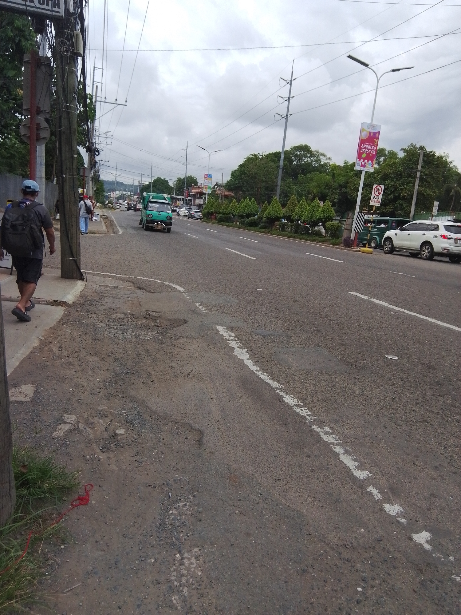 The image depicts a street scene in what appears to be Davao City, Philippines, on an overcast day. The foreground shows a paved road with visible potholes and a white dashed line marking the lane. To the left, a man wearing a blue cap and a backpack is walking on a sidewalk next to a utility pole covered in wires. Behind him, other pedestrians are also walking. In the middle ground, a bright green jeepney is driving on the road. Further down the road, other vehicles, including a white SUV, are visible.
Along the side of the road, there are trees and decorative bushes. A streetlamp with a banner hangs above the road. The banner is predominantly pink and features text in Filipino, with the words "Uli! Diay!" and "SPARKY LIFESTYLE" clearly visible. Below the banner, a "NO U-TURN" sign is posted. The sky is cloudy, suggesting a potentially humid or rainy climate. The overall scene conveys a sense of everyday urban activity in a tropical environment. A strand of red string lies on the ground near the grassy edge of the sidewalk in the lower left corner.
