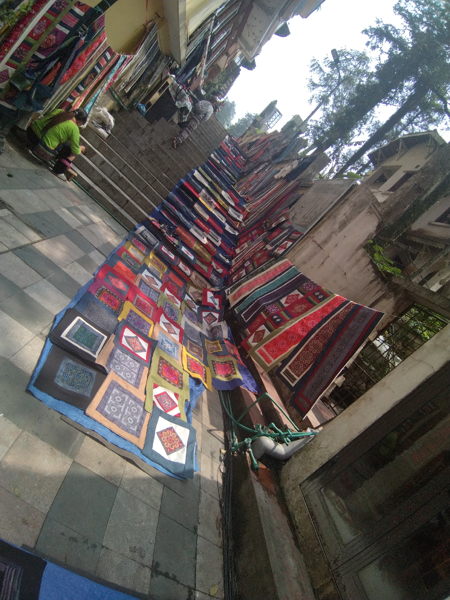 The image depicts a street scene in Sa Pa Ward, Vietnam, showcasing an outdoor marketplace. A set of concrete stairs forms the backdrop, with vendors and customers interacting along the steps. A vendor is seen crouching next to the stairs, likely arranging or selling their wares.
The primary focus is a colorful display of textiles, including carpets and smaller square fabrics, laid out across the stairs and pavement. These items exhibit various patterns and colors, indicative of local craftsmanship. Some textiles are hung along the side of the stairs, held up by ropes or other supports.
The surrounding environment consists of buildings with weathered facades, trees, and other urban elements. The perspective is slightly angled, offering a view of the street stretching upwards along the stairs. The lighting suggests daylight conditions, with shadows cast across the scene.
