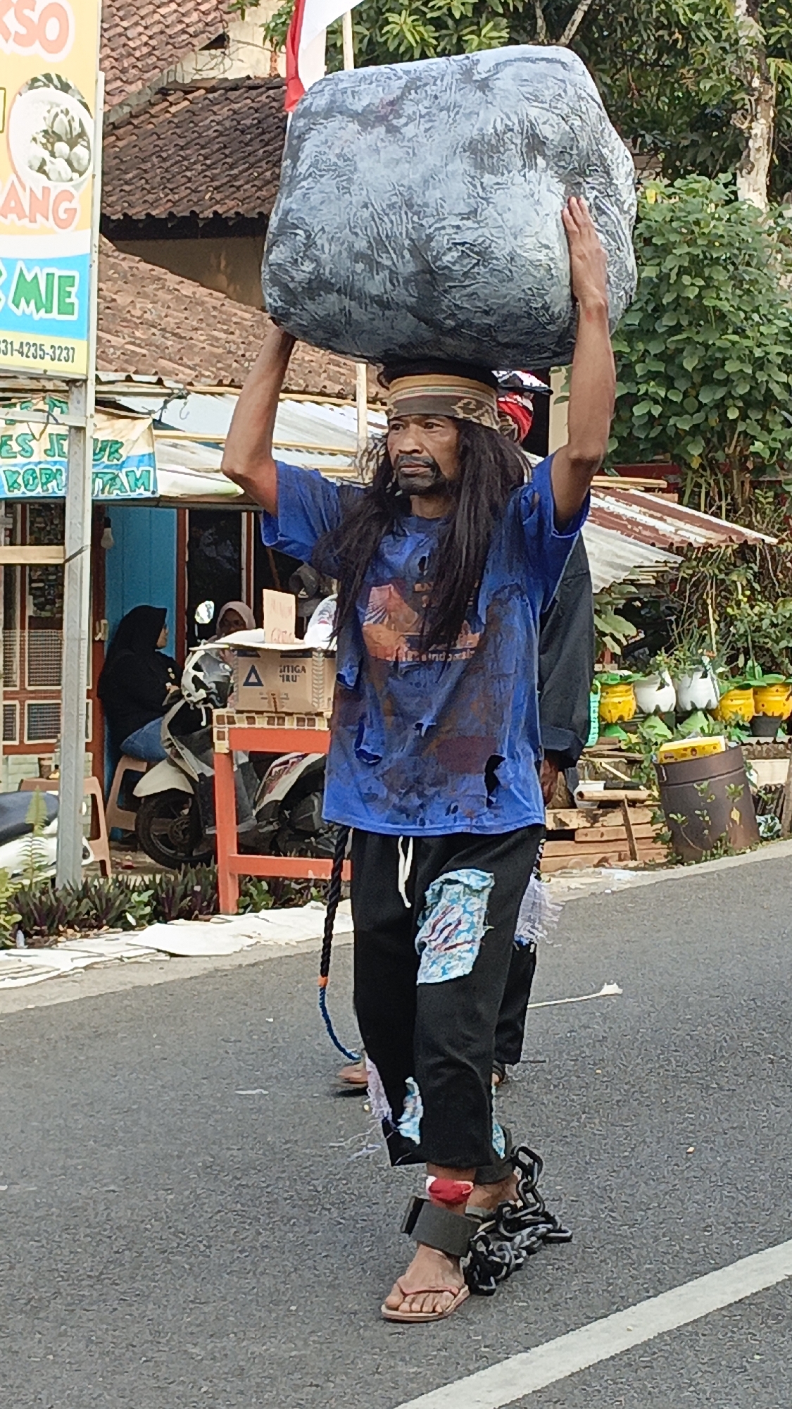 In Krajan Lor, Indonesia, a man with long, dark hair and a beard walks down a paved street, balancing a large, grey, rock-like object on his head. He is wearing a ripped blue t-shirt with a graphic design, black pants with colorful patches, and flip-flops. His ankles are bound with chains. Behind him, another person, partially visible and also in chains, follows. In the background, there are buildings with corrugated roofs, a scooter, and potted plants. A sign with text in Indonesian is visible on the left side of the image. The scene appears to be outdoors during daylight.