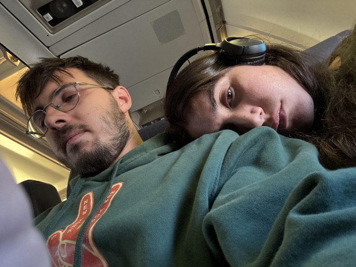 The image depicts two individuals seated within the interior cabin of an airplane; it does not show a location within the city of Tzafria, Israel. The male subject, positioned on the left and slightly to the foreground, has short dark hair, a beard, and wears round, wire-frame glasses. He is dressed in a dark green hooded sweatshirt with a faded red and white graphic on the chest, depicting a hand forming a peace sign, with the word "NEW YORK" partially visible. His head is tilted slightly, and his gaze is directed downwards. The female subject, on the right, has long dark hair and wears dark over-ear headphones. Her head is resting on the male's right shoulder/upper chest, and she is looking directly towards the viewer. The background reveals typical airplane cabin elements, including overhead panels, vents, and lighting fixtures.