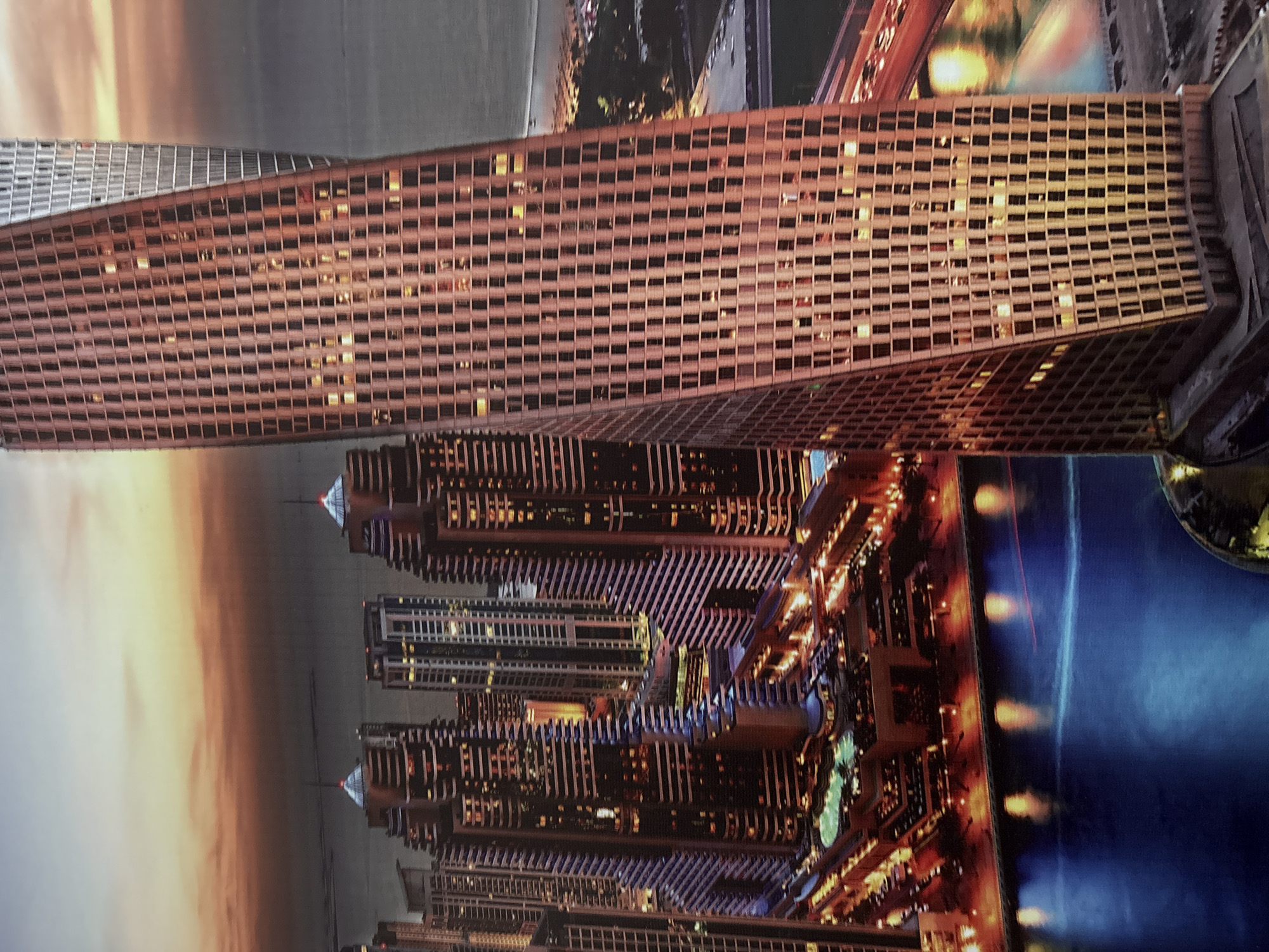 The media file presents a vibrant cityscape at twilight or night, dominated by modern architecture and artificial illumination.
In the foreground, a tall, twisting skyscraper with a distinctive grid pattern of windows stands prominently. Many of its windows are brightly lit, indicating it is evening or nighttime. The building's structure appears to twist upwards, adding a dynamic element to the urban landscape.
Behind this central building, a dense collection of other high-rise skyscrapers and buildings extends into the midground and background. These structures are also extensively illuminated, showcasing a variety of contemporary architectural designs, some featuring intricate patterns and others with pointed or pyramid-like tops.
To the right, a body of water, likely a river, canal, or bay, reflects the myriad of city lights, creating a shimmering effect. The sky above the city displays a gradient of warm hues, transitioning from orange and yellow near the horizon to softer grays higher up, characteristic of a sunset or sunrise.
No people or specific activities are visible within the frame; the focus is entirely on the architectural grandeur and the luminous urban environment. The scene evokes a sense of bustling modernity and impressive engineering. There is no visible text in the image. The high concentration of unique, modern skyscrapers suggests a location known for rapid urban development, such as Dubai.
