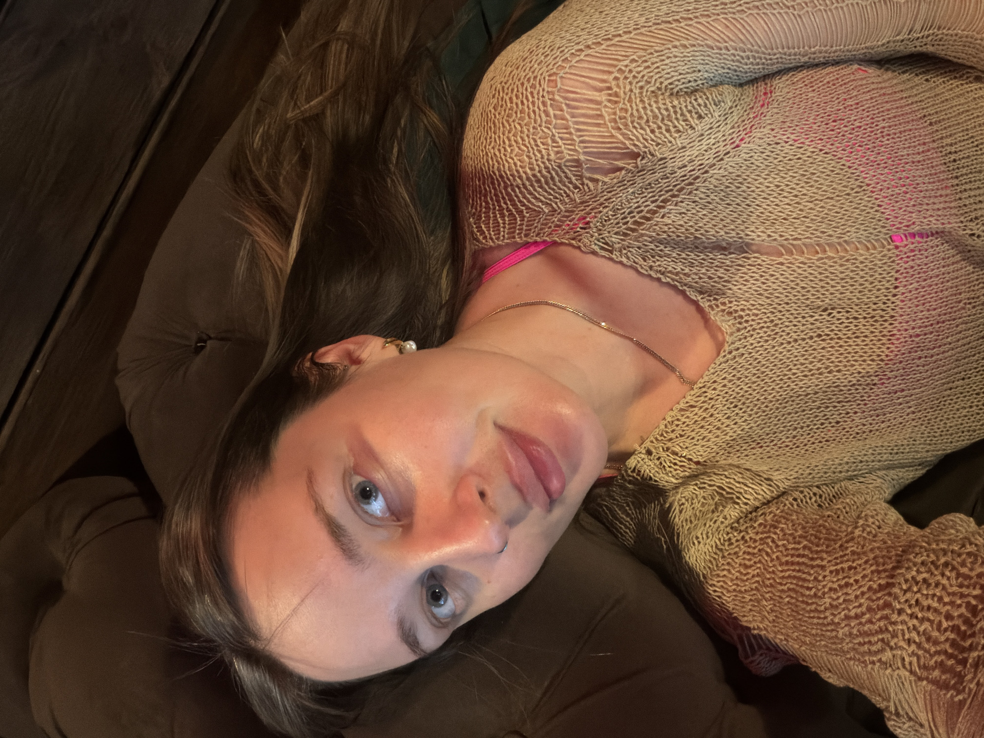A woman with long brown hair and blue eyes is lying on a brown cushion. She is wearing a beige, crocheted-style top over a pink garment, along with a gold necklace. She has a nose ring and an earring. The background is a dark, wooden surface. The location and city are not discernible.