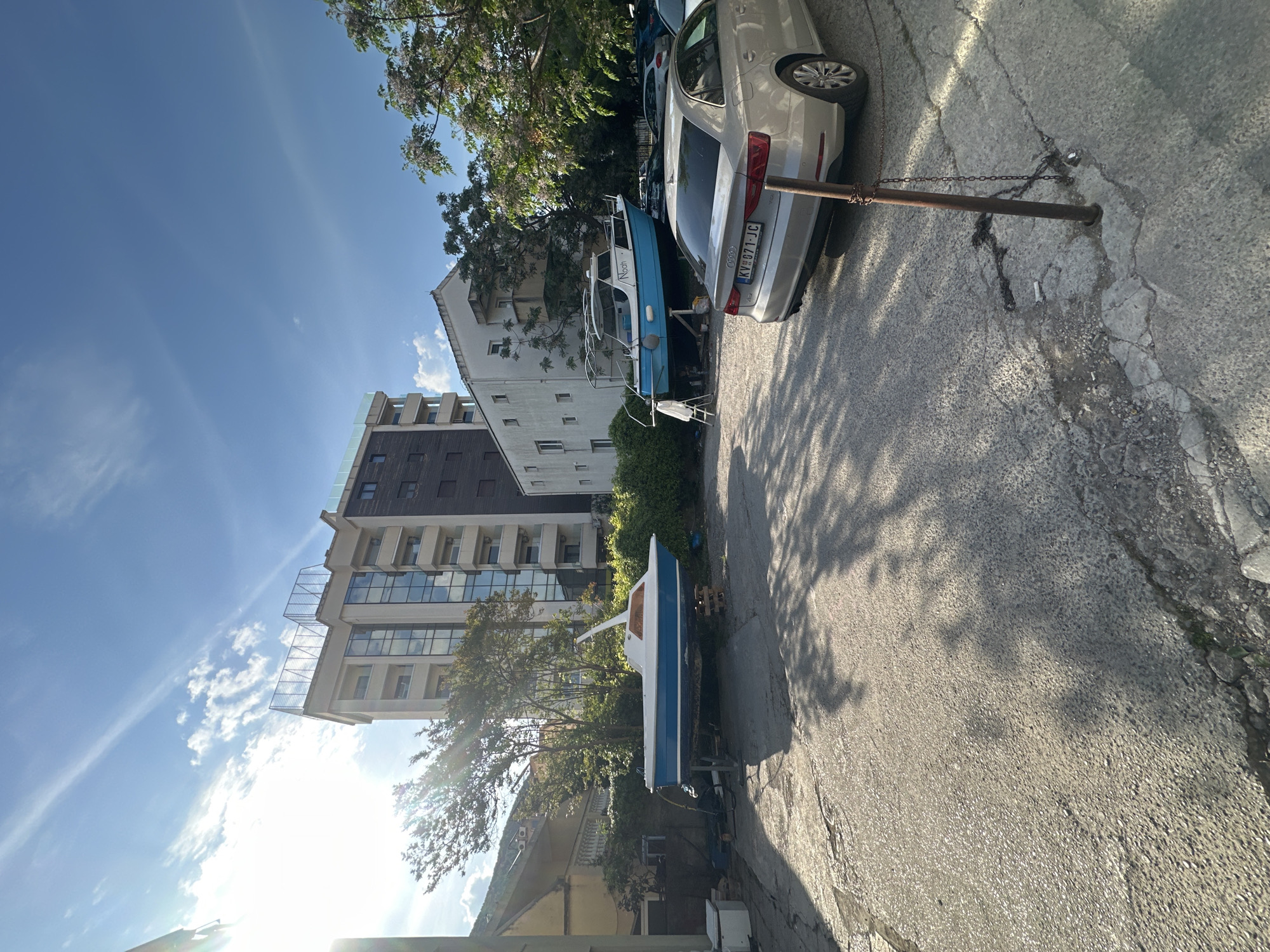 The image captures a sunny outdoor scene in Budva, Montenegro, likely during the day, with strong sunlight creating prominent shadows and lens flare on the left side of the frame. The sky is clear blue with some wispy clouds.
In the foreground, a cracked and worn asphalt road is visible. A rusty metal pole anchored in the ground, with a chain attached, stands on the right side of the road.
The midground features several parked vehicles and boats. A silver sedan, likely an Audi, is parked with its rear facing the viewer. Its license plate, "KV 071-JC," is clearly visible. Behind this car, a blue and white boat, possibly a small yacht or cabin cruiser, is resting on a trailer. The name "Nuon" or "Noon" is written on its side. Another smaller, blue and white boat is visible further down the street on the left, also appearing to be on a trailer. Green trees and dense foliage are scattered throughout the scene.
In the background, several multi-story buildings line the street. One prominent building on the left features a modern design with extensive glass windows and balconies. An older, more traditional building is situated to its right. The scene depicts a static arrangement of boats and cars alongside buildings in what appears to be a coastal or residential area.