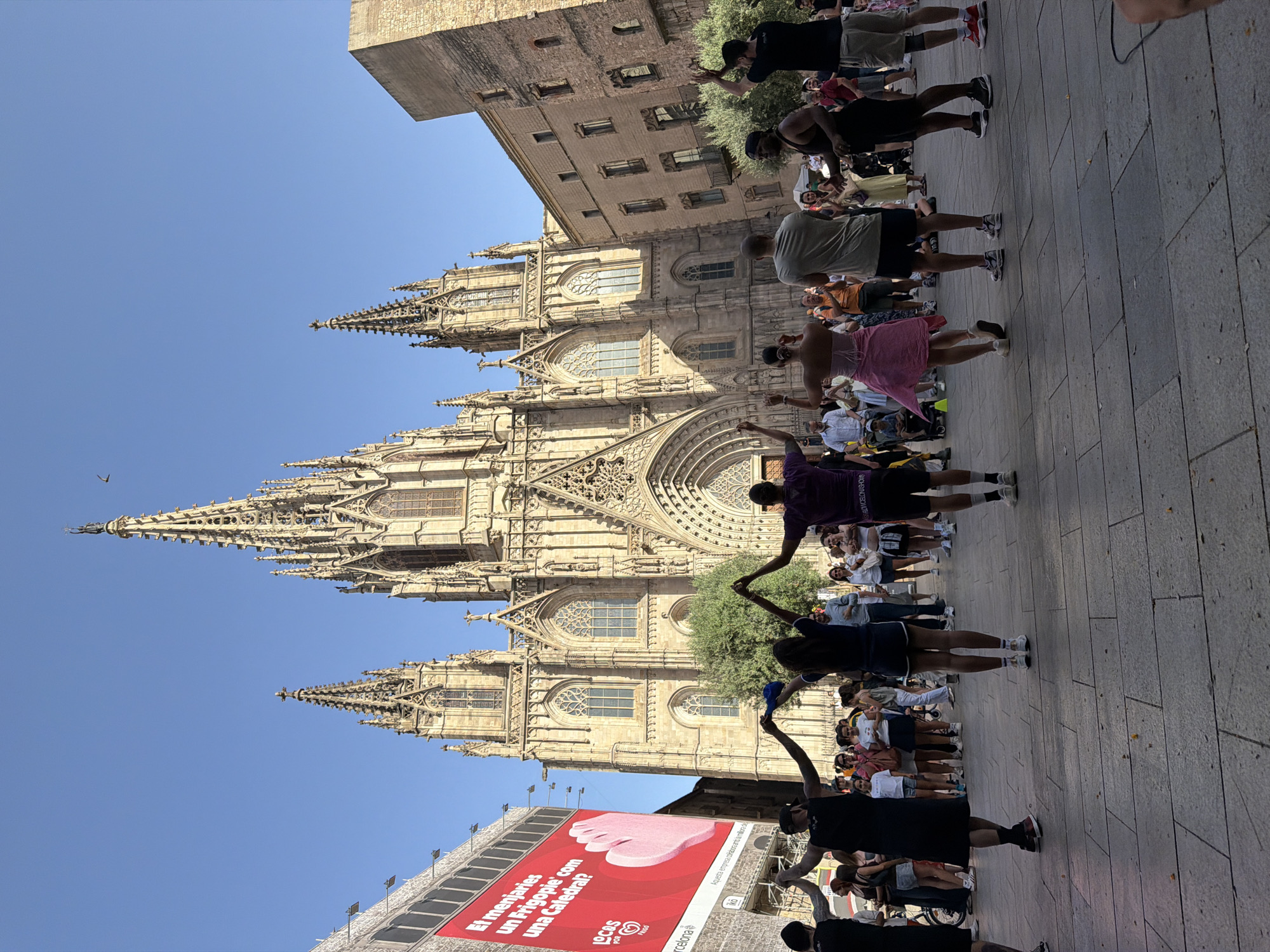 This is a photograph taken outdoors in Barcelona, Spain, featuring the Barcelona Cathedral as the prominent backdrop. The cathedral is a large, ornate Gothic structure with intricate spires, arches, and stained-glass windows. In front of the cathedral, a group of people are gathered. Some are dancing or holding hands in a circle, while others stand and watch. The foreground shows a paved plaza with a light gray stone surface. The sky above is clear and blue. To the left of the cathedral, a large billboard with red and white lettering is visible. The people in the foreground are diverse in age and attire, with some wearing casual summer clothing. The overall scene suggests a public event or gathering taking place in a historic urban setting.