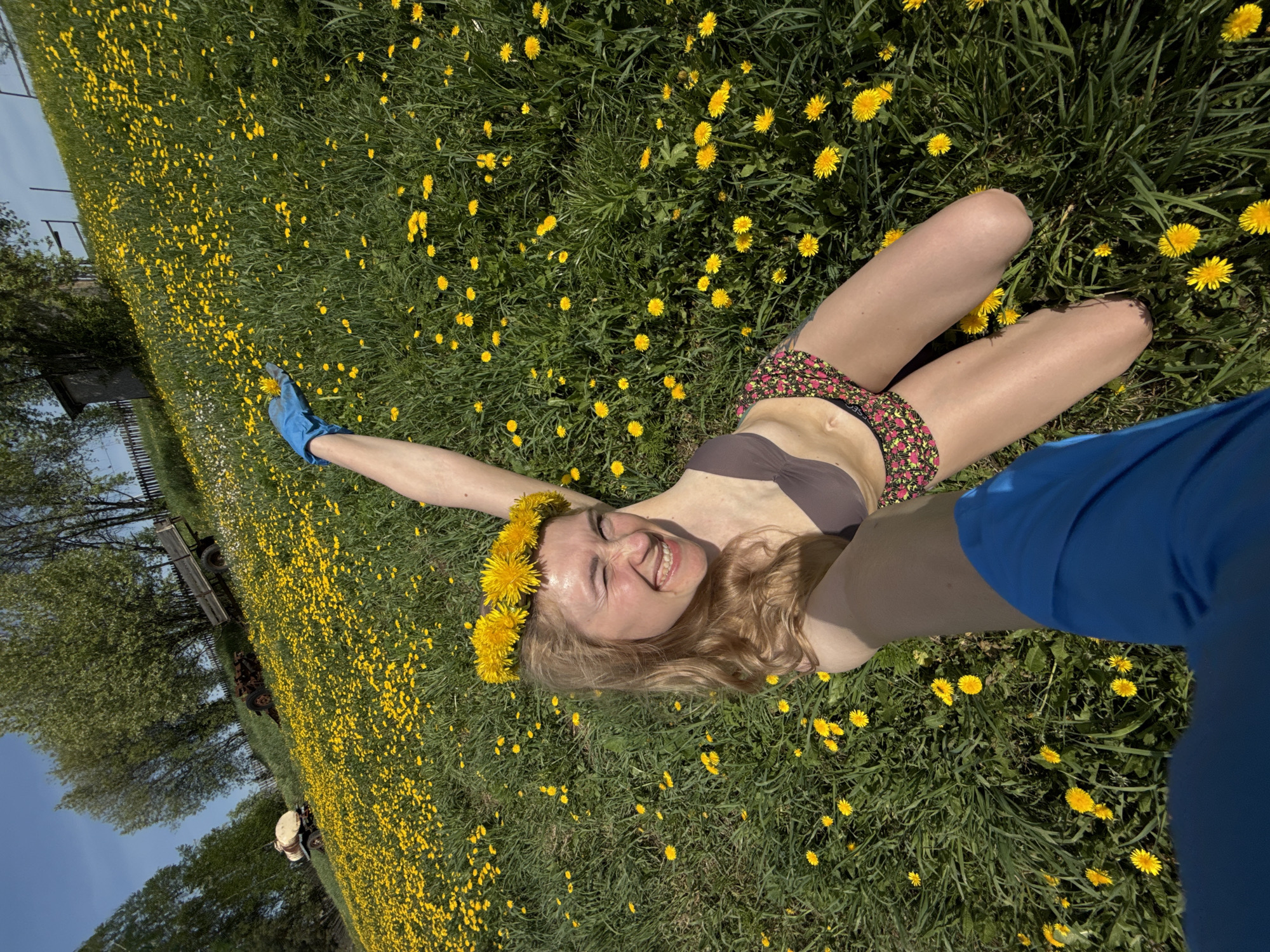A woman lies in a field of dandelions in Nizhnyaya Chermoda, Russia. She is wearing a brown bikini top and floral print bottoms, and a crown of dandelions on her head. Her blonde hair is long and wavy. She is smiling and has one arm raised in the air, wearing a blue glove. A person wearing a blue shirt is partially visible in the right side of the frame. In the background, a building, trees, and a blue sky are visible. The field is densely covered with yellow dandelions.