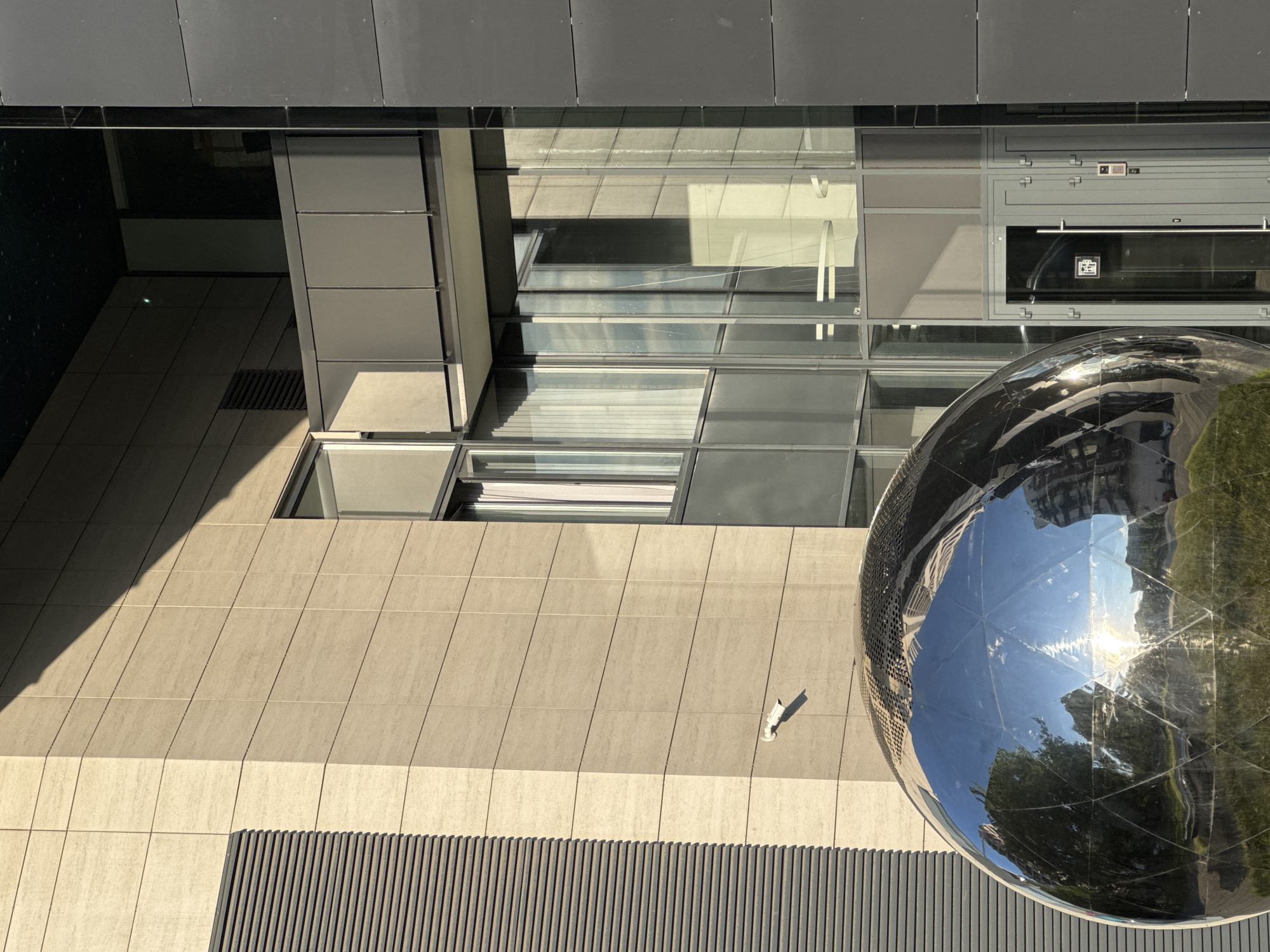 This abstract, high-angle image captures a modern architectural scene dominated by geometric forms, varied textures, and reflective surfaces. The viewpoint looks down into what appears to be a building facade or an enclosed urban courtyard.
In the upper part of the frame, dark grey metallic cladding panels are visible. Below this, a section of large glass windows reflects elements of the surrounding environment, including what appears to be a lighter-colored ceiling or wall and indistinct architectural shapes. Within one of these reflective glass panels, a small, square logo or sign is visible, featuring indecipherable text or symbols against a dark background, possibly indicating a unit or business.
The central and lower-left areas are characterized by a light beige or tan tiled wall, composed of rectangular panels with vertical grout lines. A security camera, small and white, is mounted on this wall, pointing slightly downward. Below the tiled wall, a section of dark, vertically slatted material, possibly a ventilation grate or decorative paneling, runs horizontally across the bottom.
A large, highly reflective metallic sphere, likely a piece of modern sculpture or an architectural feature, occupies the lower right portion of the image. Its segmented, geodesic-like surface brilliantly reflects the bright sky, strong sunlight, lush green trees, and the surrounding buildings.
The vivid reflections and crisp shadows indicate that the photograph was taken during the daytime, under sunny and clear weather conditions. The image conveys a sense of contemporary urban design, focusing on the interplay of light, shadow, and materiality in a static, architectural setting. There are no people or specific events occurring.