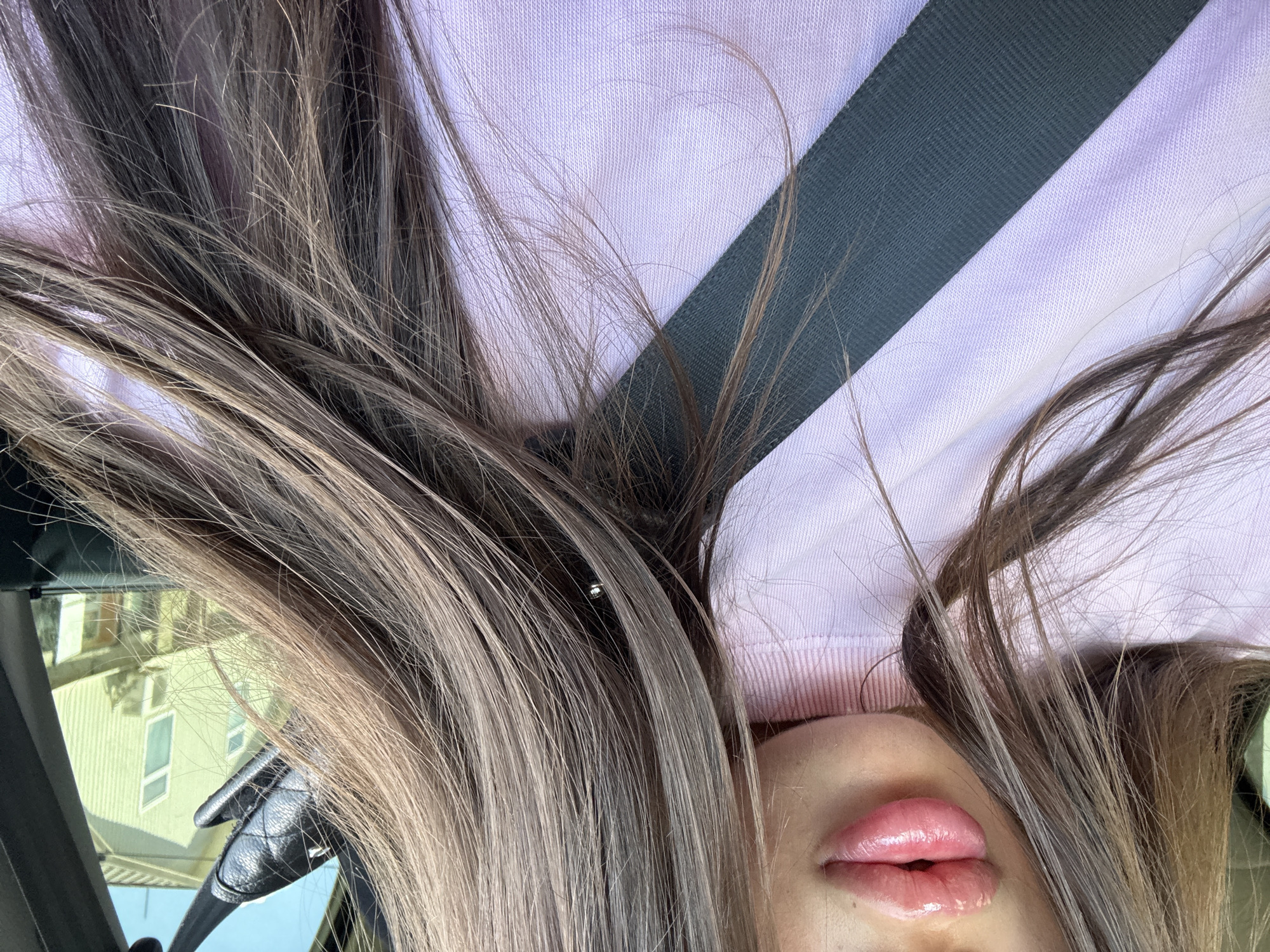 An extreme close-up shot focuses on a person's lips and part of their face, framed by their long, straight, brown hair. The lips are plump and shiny, with a pink, glossy finish. The person is wearing a light pink, high-necked sweater, over which a dark gray seatbelt is diagonally positioned. The background is blurred but suggests an outdoor environment with a building and sky visible through a window, potentially from inside a vehicle. The overall impression is a personal selfie taken in transit. The location is identified as Basarabeasca, Moldova.