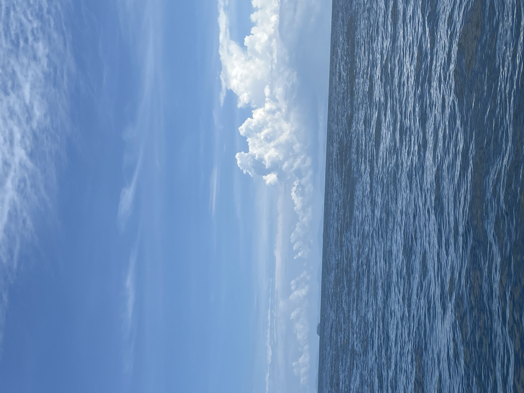 A serene seascape dominates the image, depicting the expanse of the ocean under a vast, partly cloudy sky. The water, a deep blue, is gently rippled, suggesting a calm day at sea. The horizon line is clear, separating the ocean from the sky. A chain of fluffy white clouds stretches across the upper portion of the scene, with a particularly large and dramatic cumulus cloud formation to the right, casting subtle shadows. In the far distance, a faint outline of land, possibly islands or a coastline, is visible as a dark, hazy shape against the lighter sky. The overall impression is one of peacefulness and natural beauty. The scene is likely Haad Yao, Thailand, known for its tranquil waters and picturesque coastal views.