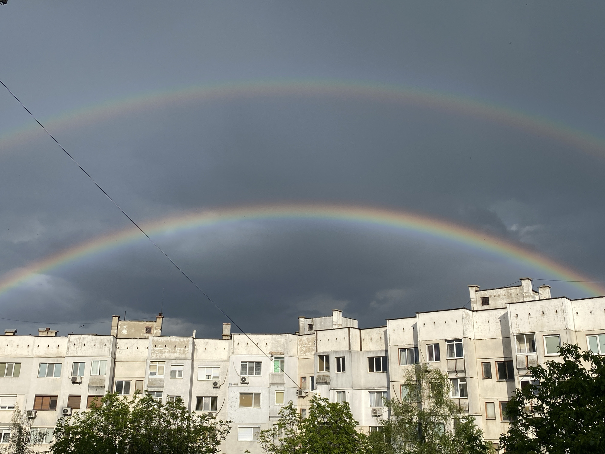 The image shows a double rainbow in the sky over a residential building. The building is a multi-story apartment complex with a light-colored facade and multiple windows, some of which have air conditioning units installed. Trees with green foliage are visible in the foreground, partially obscuring the base of the building. An overhead wire stretches across the sky in the upper left portion of the image. The sky is overcast with dark, stormy clouds, contrasting with the bright colors of the rainbows. The architecture of the building, the style of windows, and the presence of air conditioning units suggest an urban setting in Eastern Europe. Based on the building style and architecture, this scene is consistent with the city of Pleven, Bulgaria. There is no people or actions visible in the picture.