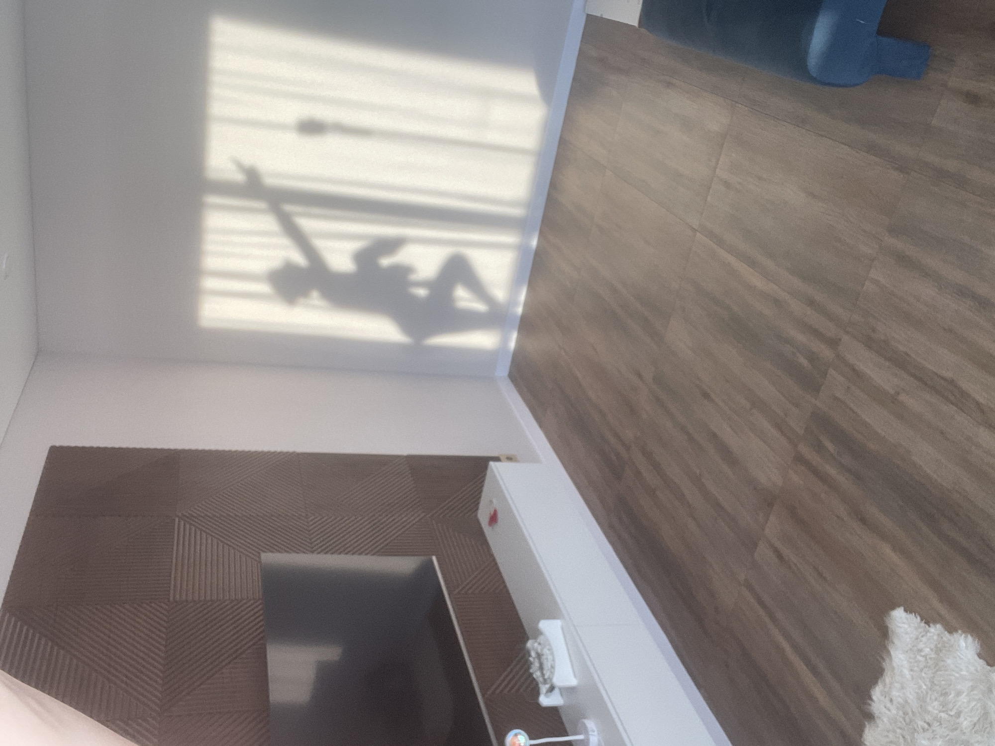 The image captures an indoor room with distinct areas defined by different wall and floor coverings. Dominating the upper left quadrant is a bright white wall, illuminated by direct sunlight streaming through a window, casting prominent shadows. The most notable shadow is that of a person, likely female, depicted in a reclining or playful pose. One arm is raised with two fingers extended, while the other arm is bent, and the legs are crossed. Overlapping the person's shadow are horizontal lines from window blinds, indicating the source of the light.
To the right of the white wall, a section of light brown wooden floor tiles is visible, continuing towards the upper right corner. In the top right, a dark blue piece of furniture, possibly a couch or chair, is partially seen. The lower left portion of the room features a dark brown wall panel with a geometric, textured pattern, suggesting a decorative feature or a media wall. A dark, rectangular screen, likely a television, is partially visible against this panel.
Running along the bottom right side, adjacent to the textured panel and extending towards the wooden floor, is a long, white low-profile cabinet or shelf unit. On this unit, a clear, faceted decorative object (possibly crystal or glass) is visible, along with a small, colorful item beneath it. A patch of fluffy white material, likely a rug, is seen in the bottom right corner, partially on the wooden floor.
The strong, crisp shadows indicate a sunny day, likely morning or late afternoon, when the sun's angle is lower. The overall impression is that of a modern, well-lit interior space. No text is visible in the image. The setting is an interior room in Podshivalovo, Russia.