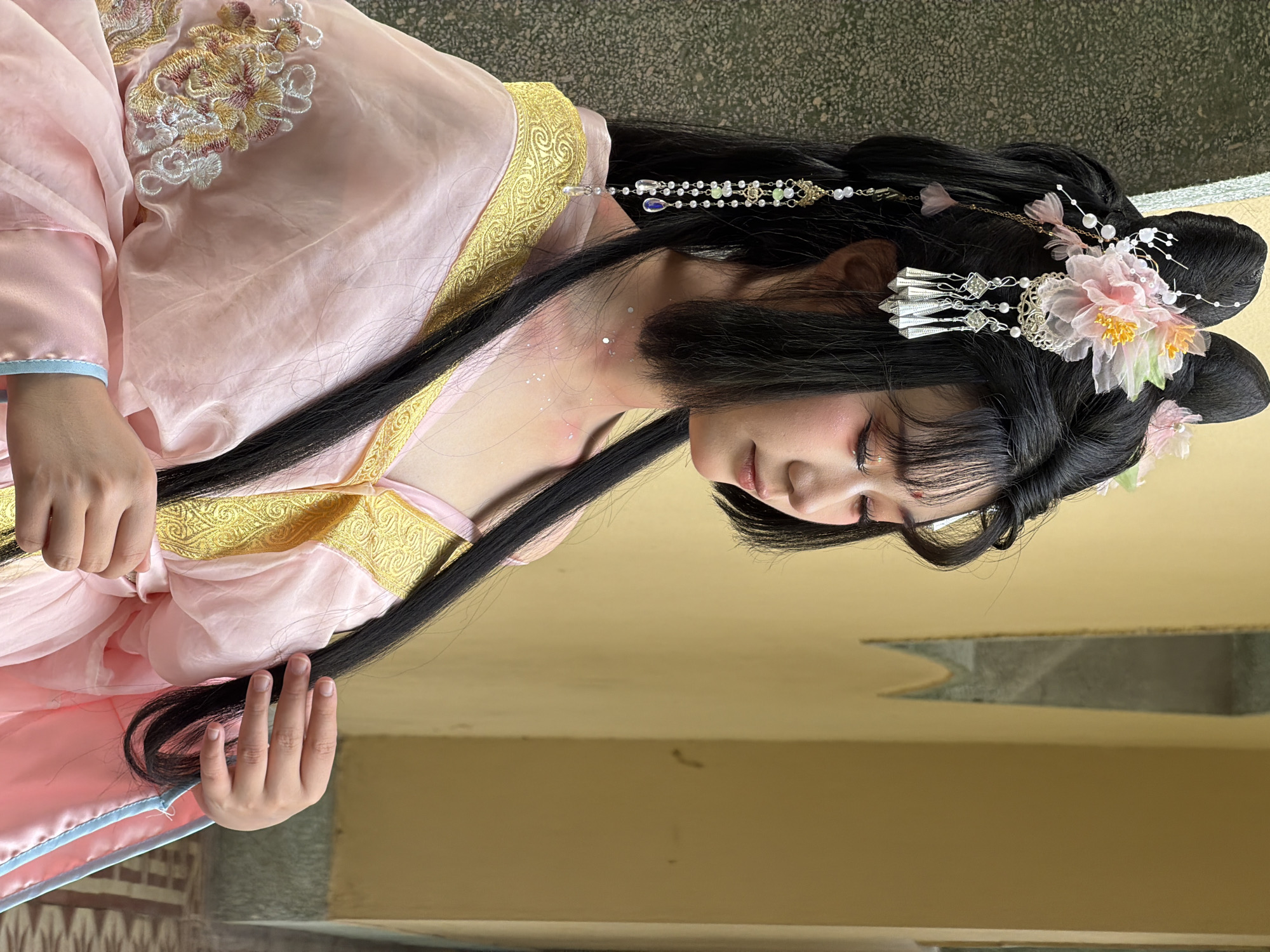 A young East Asian woman with long black hair styled in an elaborate updo adorned with pink flowers and dangling silver ornaments poses in front of a textured green wall and a yellow wall. She is wearing a pink traditional Chinese garment with gold embroidery on the collar and sleeve. Her makeup includes pink eyeshadow and glitter on her collarbone. Her eyes are closed as she gently touches her hair. The background appears to be an outdoor setting with architectural elements. The flooring is a mosaic of brown and white tiles. The scene evokes a sense of traditional Chinese culture and aesthetics.