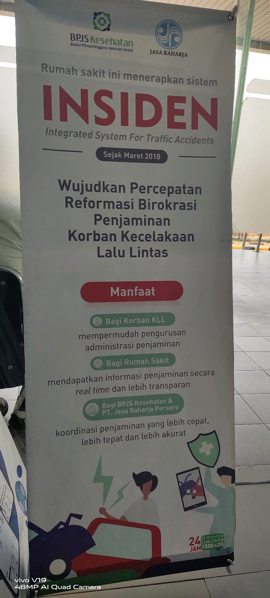 This is a banner displayed outdoors, likely in a public space in Pekanbaru, Indonesia, possibly at a government office or hospital. The banner promotes a system called INSIDEN, which stands for "Integrated System For Traffic Accidents." The banner features logos for BPJS Kesehatan and Jasa Raharja, both Indonesian government agencies related to health and social security. The text on the banner is in Indonesian and details the benefits of the INSIDEN system for victims of traffic accidents, hospitals, and the coordinating agencies. The lower part of the banner includes a graphic illustration depicting a car accident scene with people, a mobile phone displaying a shield with a bug, and a 24-hour care center contact number. The bottom left corner shows the text "vivo V19 48MP AI Quad Camera." The overall environment appears to be paved with tiles, and a building or structure is visible in the background.