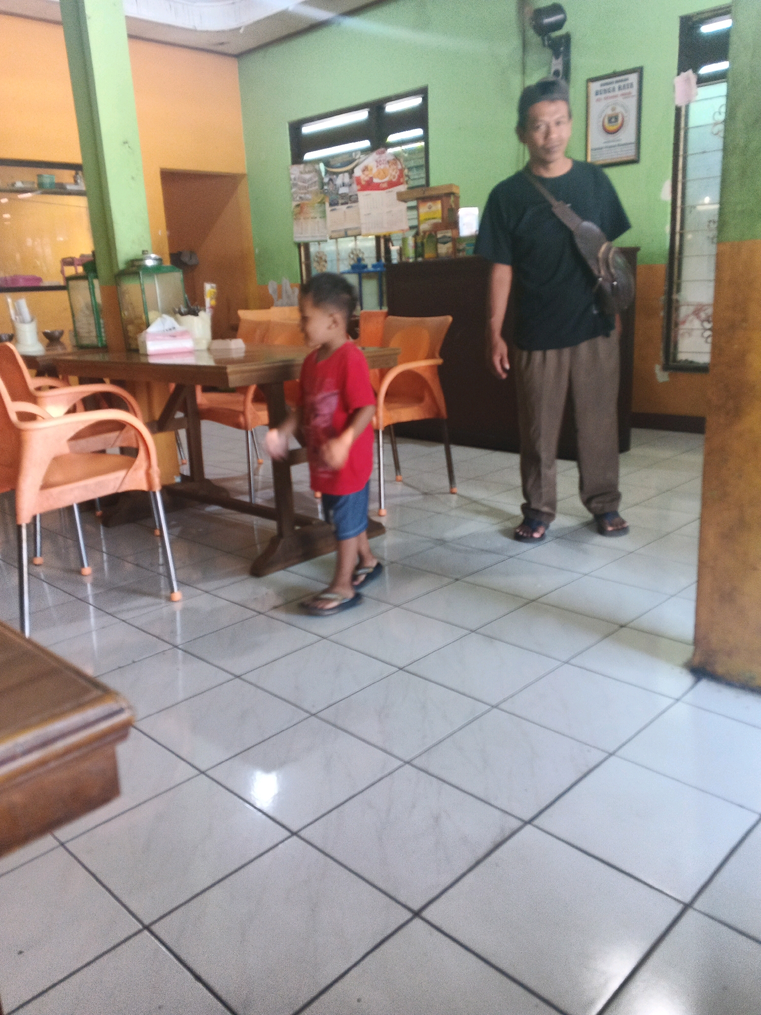 The image depicts an indoor scene, likely a restaurant or cafe, in Ungaran, Indonesia. A young boy in a red t-shirt and blue shorts is standing in the foreground, facing away from the camera and slightly to the left. He is wearing dark sandals. To his right and slightly further back, an adult male in a dark t-shirt and brown pants is standing, facing the camera. He has a dark bag slung across his shoulder and is also wearing sandals. The environment includes orange chairs and a wooden table in the middle ground. The walls are painted in green and orange hues, and there are windows and framed pictures visible. The floor is tiled with white squares.