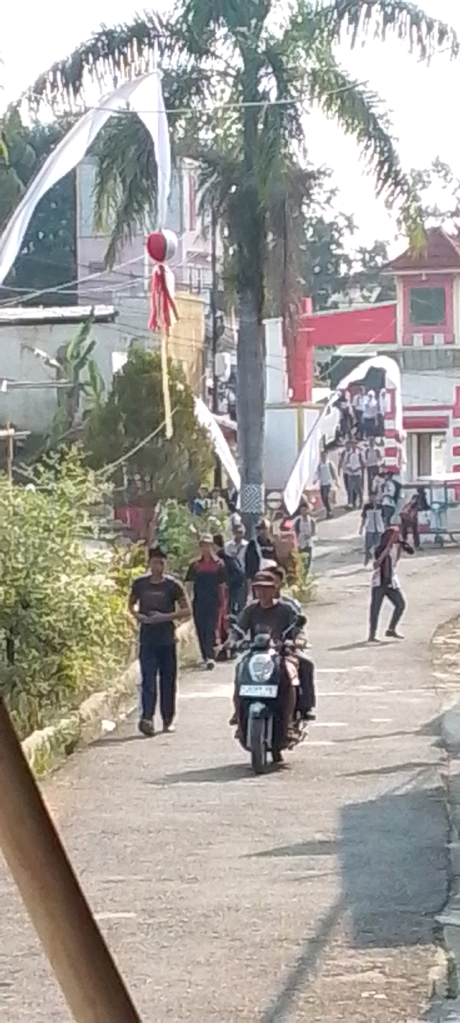 The image depicts a street scene in Cilaku, Indonesia, likely during an event. A person is riding a scooter, navigating a paved road lined with greenery on the left and buildings in the background. Several other individuals are visible, some walking and others standing. Decorative white and red banners are strung across the scene, with palm trees and other vegetation contributing to the tropical environment. The overall atmosphere suggests a public gathering or celebration.