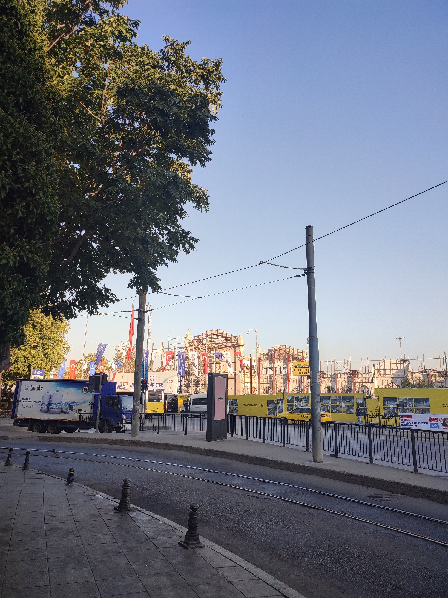 An urban street scene in Kadıköy, Turkey, features a sidewalk with ornate bollards bordering a road with tram tracks. On the right side of the road, a black metal fence encloses an area with yellow banners and scaffolding, indicating construction or renovation. Several yellow vehicles, including what appears to be an Uber taxi, are visible behind the fence. A blue truck with a "Galata" advertisement on its side is parked on the left side of the road, near a large tree with green foliage. Overhead, tram wires are strung across the sky, which is clear and blue. In the background, flags are displayed on poles, and parts of buildings, some under construction, are visible. The lighting suggests it is daytime, possibly late afternoon or early morning due to the long shadows.