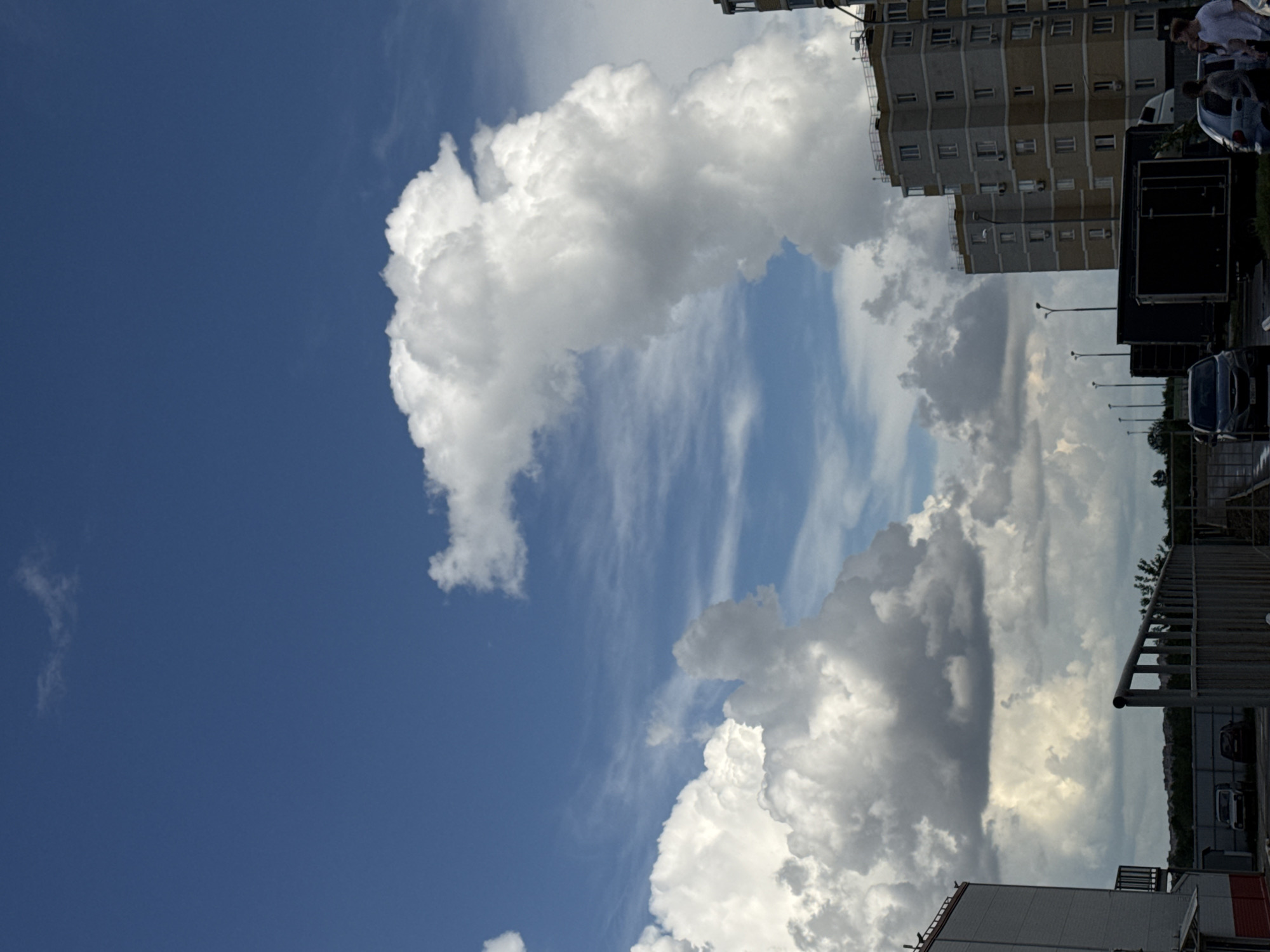 The image shows a partly cloudy sky over Krasnodar, Russia. A tall apartment building is visible on the right, next to a parking lot. Several cars, street lamps, and people are observed near the building. In the foreground, parts of industrial structures can be seen. The sky is primarily blue with large, white cumulonimbus clouds.
