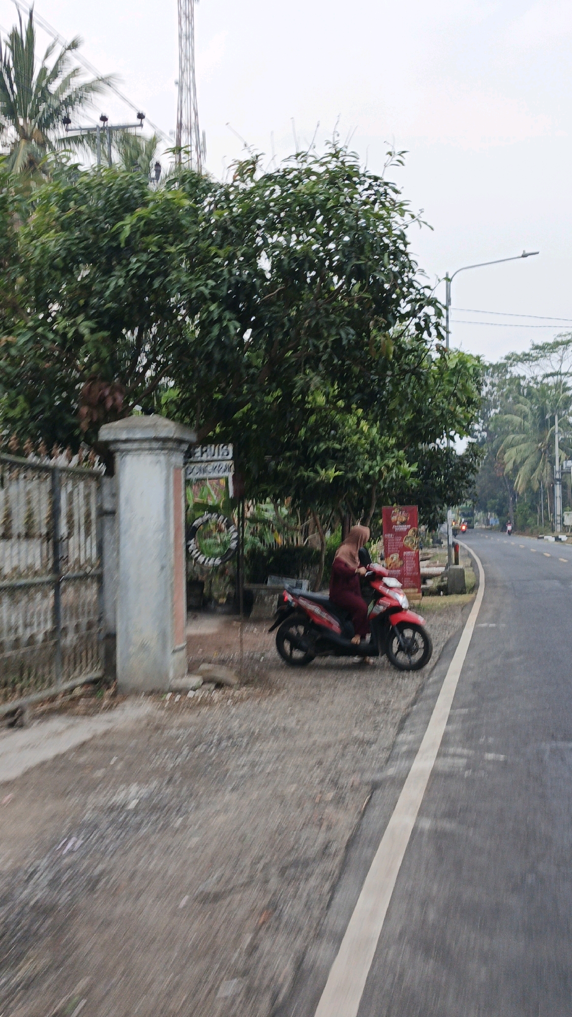 The image depicts a scene in Sukaresik, Indonesia, focusing on a woman riding a red scooter on the side of a paved road. The woman is wearing a maroon hijab and matching clothing. She is positioned in the right half of the frame, on the shoulder of the road, which is bordered by a white line. To her left, a gate made of vertical metal bars is visible, with a thick concrete pillar in front of it. Behind the gate, lush green trees and vegetation are abundant, with a tall communication tower rising in the background. Several signs are also present in the background, some with text, including one that appears to say "ERVIS" and another with red text. The road itself appears to be a two-lane street with other vehicles, including another scooter and a car, visible in the distance. The environment suggests a semi-urban or rural setting with a mix of natural elements and infrastructure.