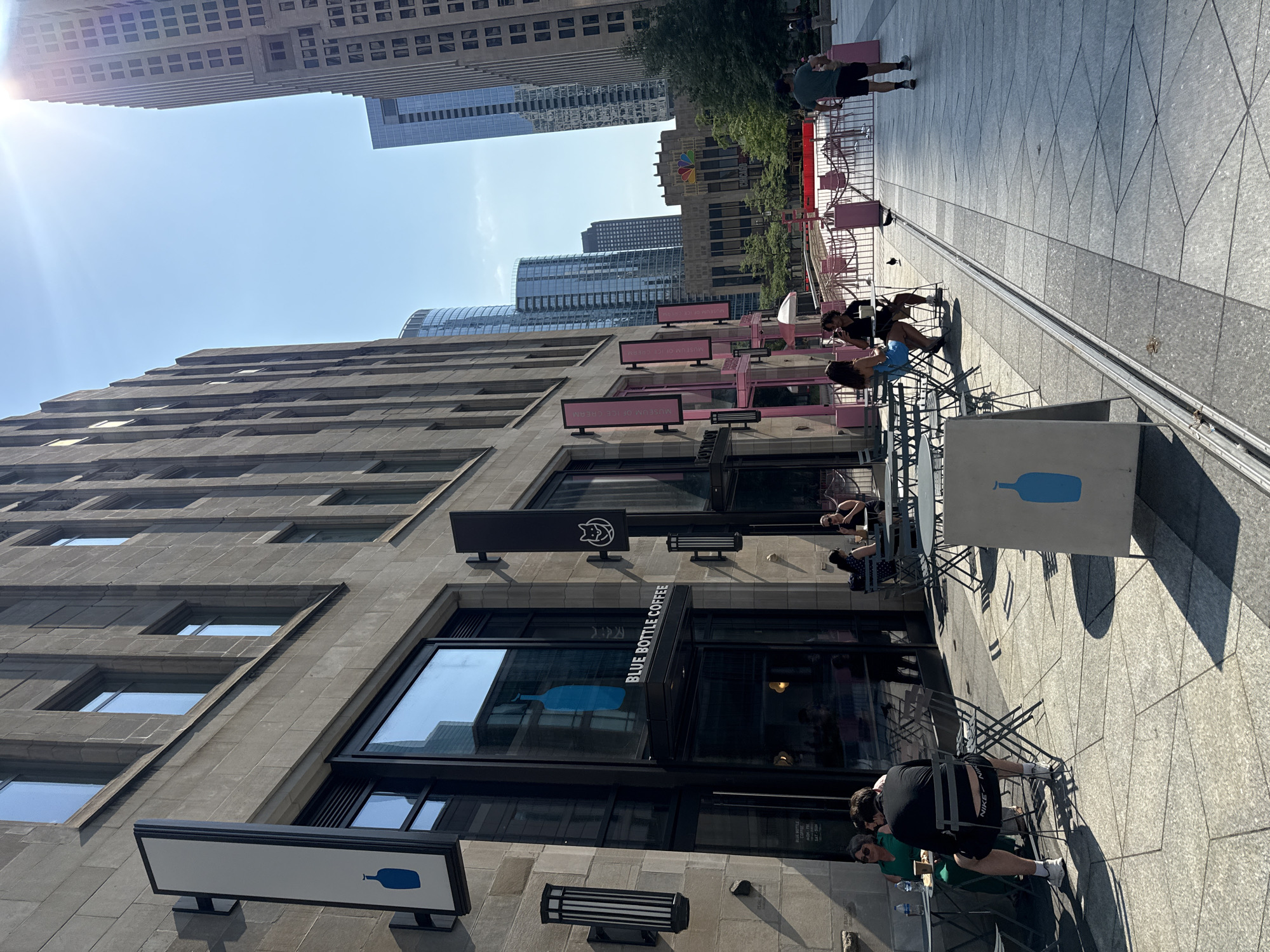 The image depicts a street scene in Chicago, Illinois, USA, on a sunny day. In the foreground, a Blue Bottle Coffee shop is visible with patrons seated at outdoor tables. The shop's facade features large glass windows and signage with the brand's distinctive blue bottle logo. Beyond the coffee shop, a row of modern skyscrapers rises into the clear blue sky, some reflecting the sunlight. In the middle ground, a fenced-off area with pink barriers suggests a temporary event or setup. A person in a green t-shirt and black shorts walks away from the camera towards the right side of the frame. The street and sidewalk are paved with geometric stone tiles, and shadows from the buildings and awnings are cast across the scene. The overall atmosphere is urban and bustling, capturing a typical summer day in the city.