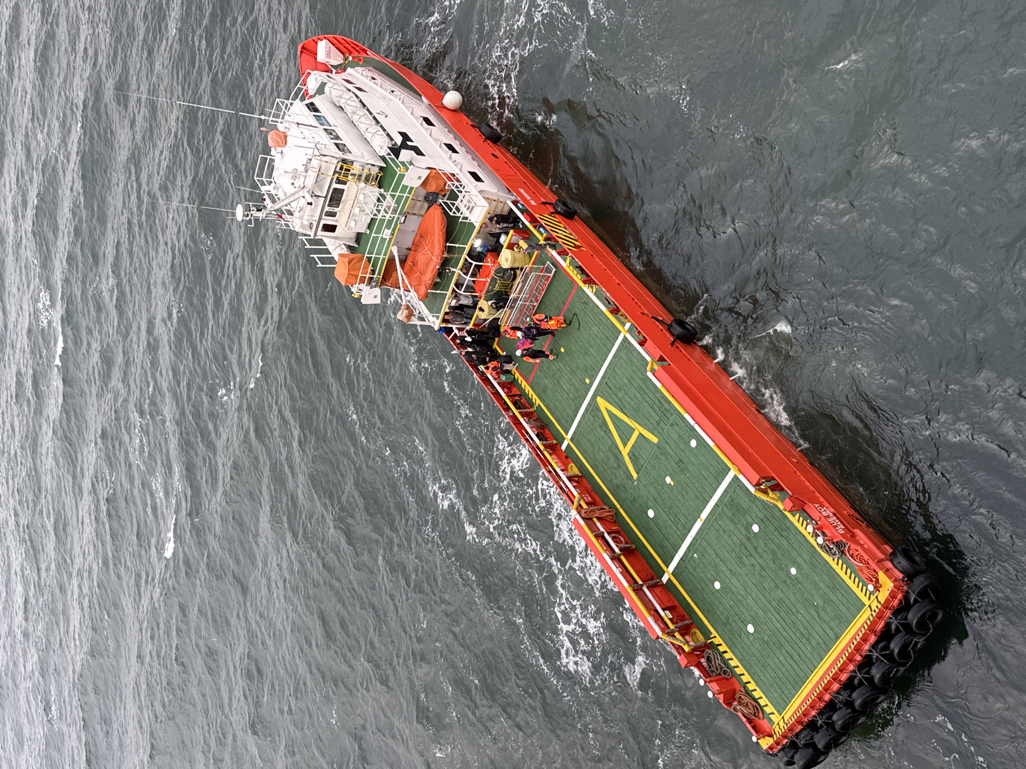 Aerial view of a red and white vessel in the ocean, likely a supply or support vessel for offshore operations. The vessel has a green helipad marked with a yellow "A". Some individuals are visible on the vessel's deck, dressed in work attire. The sea state appears moderate, with visible waves and choppy water. The location and specific city in Italy are not identifiable from the image.