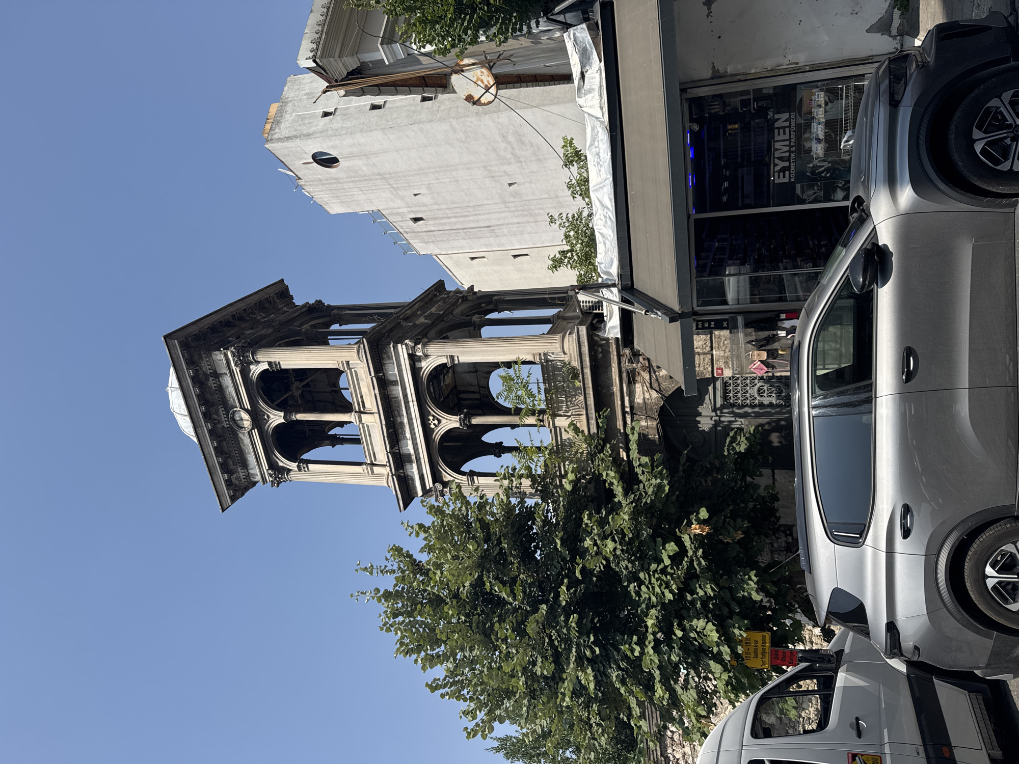 An exterior, eye-level, wide shot captures an old stone tower with arched openings and a dome, situated in an urban setting in Istanbul, Turkey. The tower, adorned with classical architectural elements like columns and cornices, appears weathered and partially ruined, with some greenery growing on its balconies. To the right of the tower, a stark white, modern building with a circular window and several smaller rectangular windows rises. In the foreground, a grey van is partially visible on the left, and a silver SUV is parked on the right, with its rear end facing the viewer. In front of the SUV, a storefront with a dark awning displays the text "EYMEN KOZMETİK & PARFÜMERİ" (Eymen Cosmetics & Perfumery). A mature, leafy tree with dense foliage stands prominently between the tower and the van. The sky is clear and blue, indicating a sunny day. The scene depicts a blend of historical architecture and contemporary city life, with traffic and commercial activity.