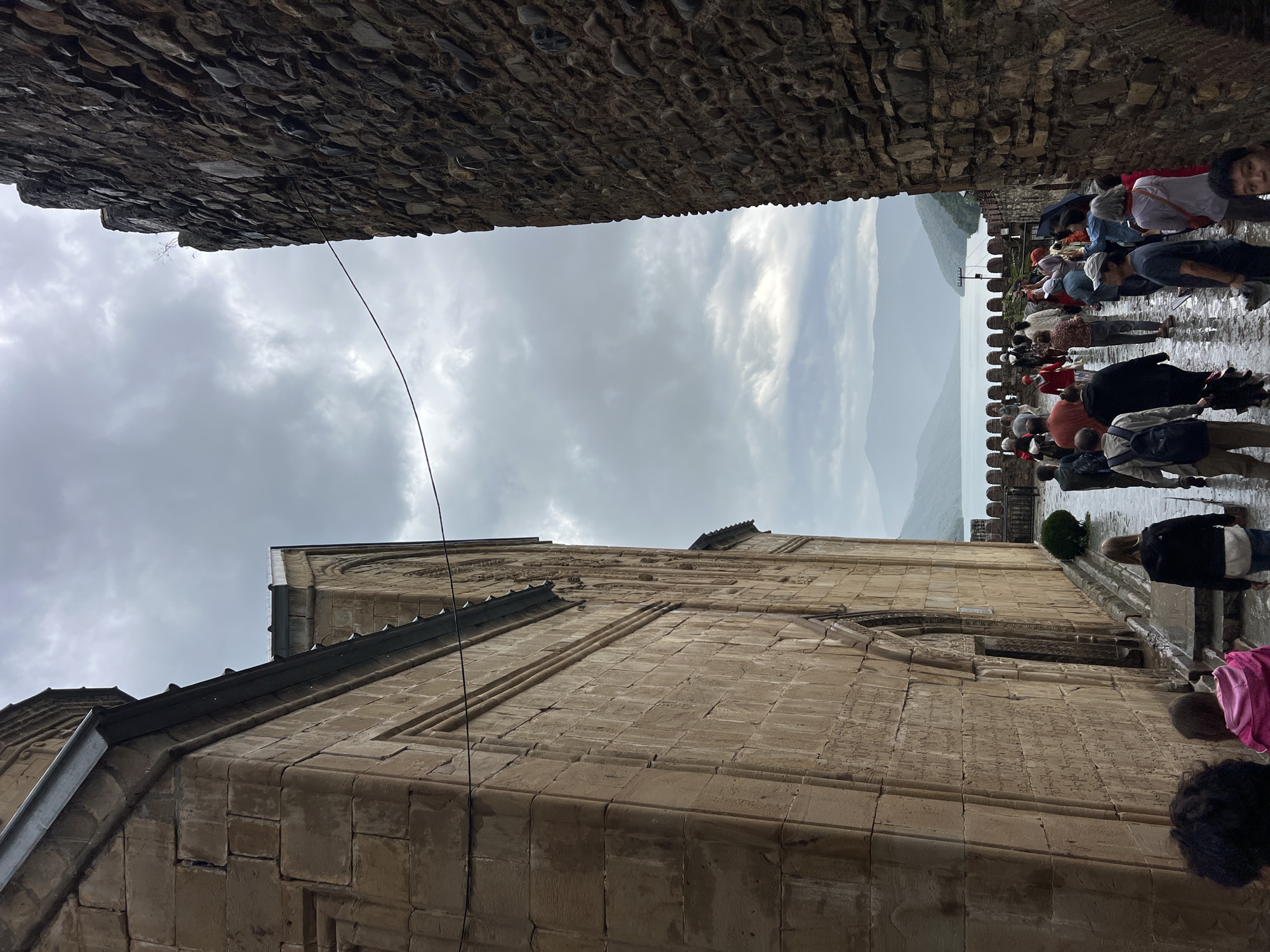 The image depicts a scene at the Ananuri fortress complex in Shalikiantkari, Georgia, under a cloudy sky. In the foreground, a path is wet, suggesting recent rain. On the left, a large stone building, part of the fortress, is visible, with its textured walls and architectural details. A narrow, shadowed passageway formed by this building and a rugged stone wall on the right leads the viewer's eye towards a body of water and distant hills. A group of people, appearing to be tourists, are walking along the path and through the passageway. They are dressed in casual clothing, and some are holding umbrellas. The water in the background is a calm blue, bordered by a crenelated wall and a line of trees. The overall atmosphere is somber, due to the overcast weather and the ancient, imposing architecture.