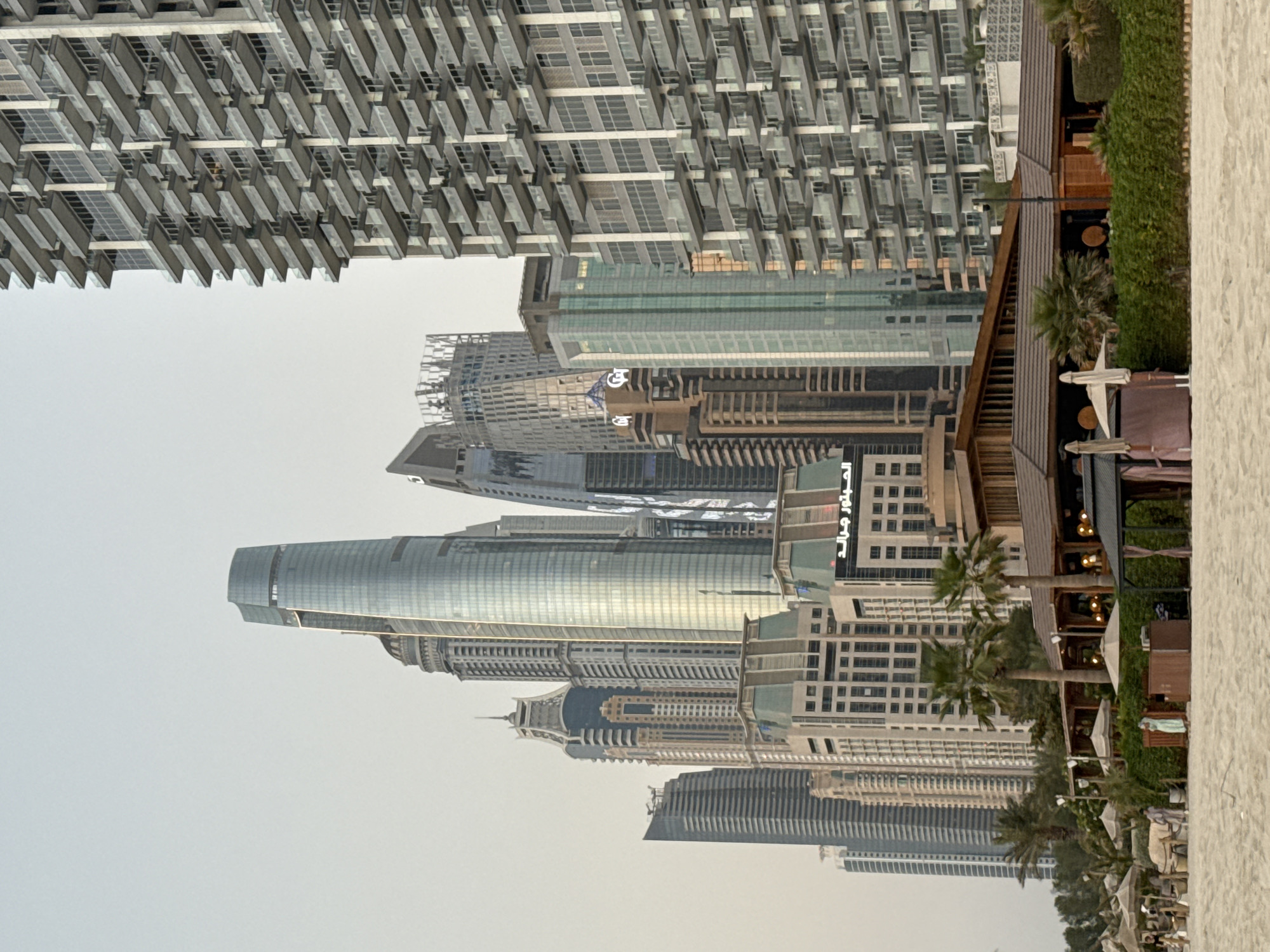 The image depicts a coastal view of Dubai Marina, United Arab Emirates. In the foreground, a sandy beach is visible with several beach umbrellas and seating areas, suggesting a resort or dining establishment. Lush green bushes and palm trees line the area behind the beach. Dominating the background is a cluster of modern skyscrapers, characteristic of Dubai's futuristic architecture. One prominent skyscraper, tall and cylindrical with a glass facade, stands out. To the right, a massive residential building with numerous balconies is featured. The sky is overcast, with a pale, hazy hue.