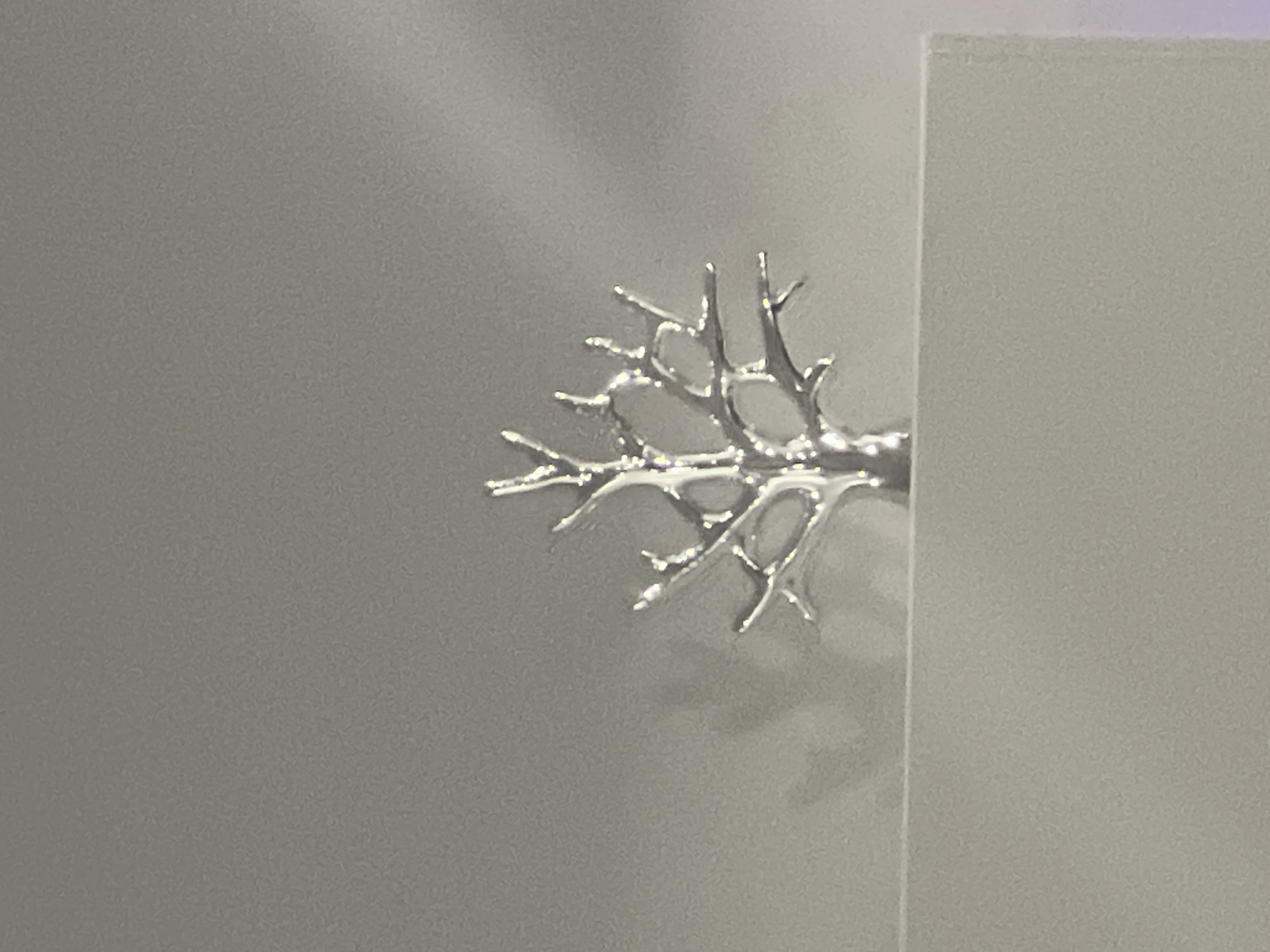 A silver metallic tree sculpture with intricate branches stands on a white surface. The sculpture is positioned in the center of the frame, casting a shadow on the light gray background behind it. The background appears to be a wall or a curtain, with subtle lighting creating variations in tone and creating a blurred effect. There are no people or animals visible in the image. The environment is minimalist and appears to be indoors. The image does not provide any information related to Podgumer, Bulgaria.