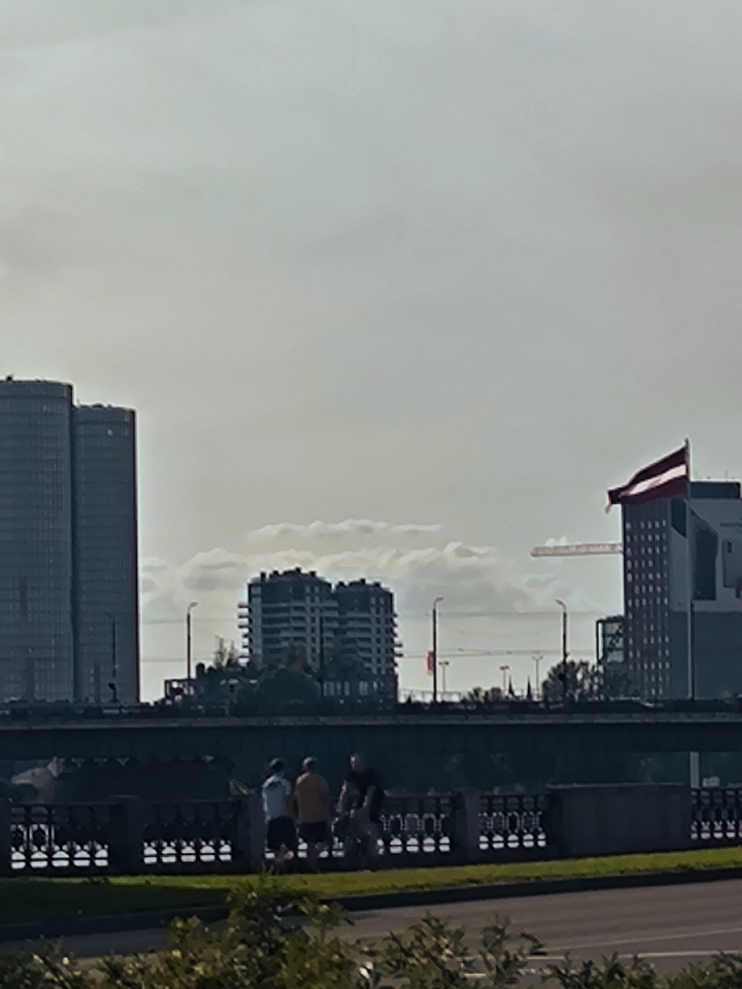 The image shows a panoramic view of the city of Riga, Latvia. In the foreground, a paved road with green grass borders can be seen. A dark, ornate metal fence runs along the top of a embankment, with a row of what appear to be decorative lights or carvings along its base. Behind the fence, four individuals are visible, standing and possibly conversing. The middle ground features a bridge spanning a body of water, with more decorative elements on its railing. In the background, several buildings rise into the sky. To the left, two tall, dark, cylindrical buildings are prominent. In the center, two shorter, rectangular apartment buildings stand. To the right, a building with a Latvian flag flying from its corner is visible, alongside a construction crane. The sky is overcast with clouds.