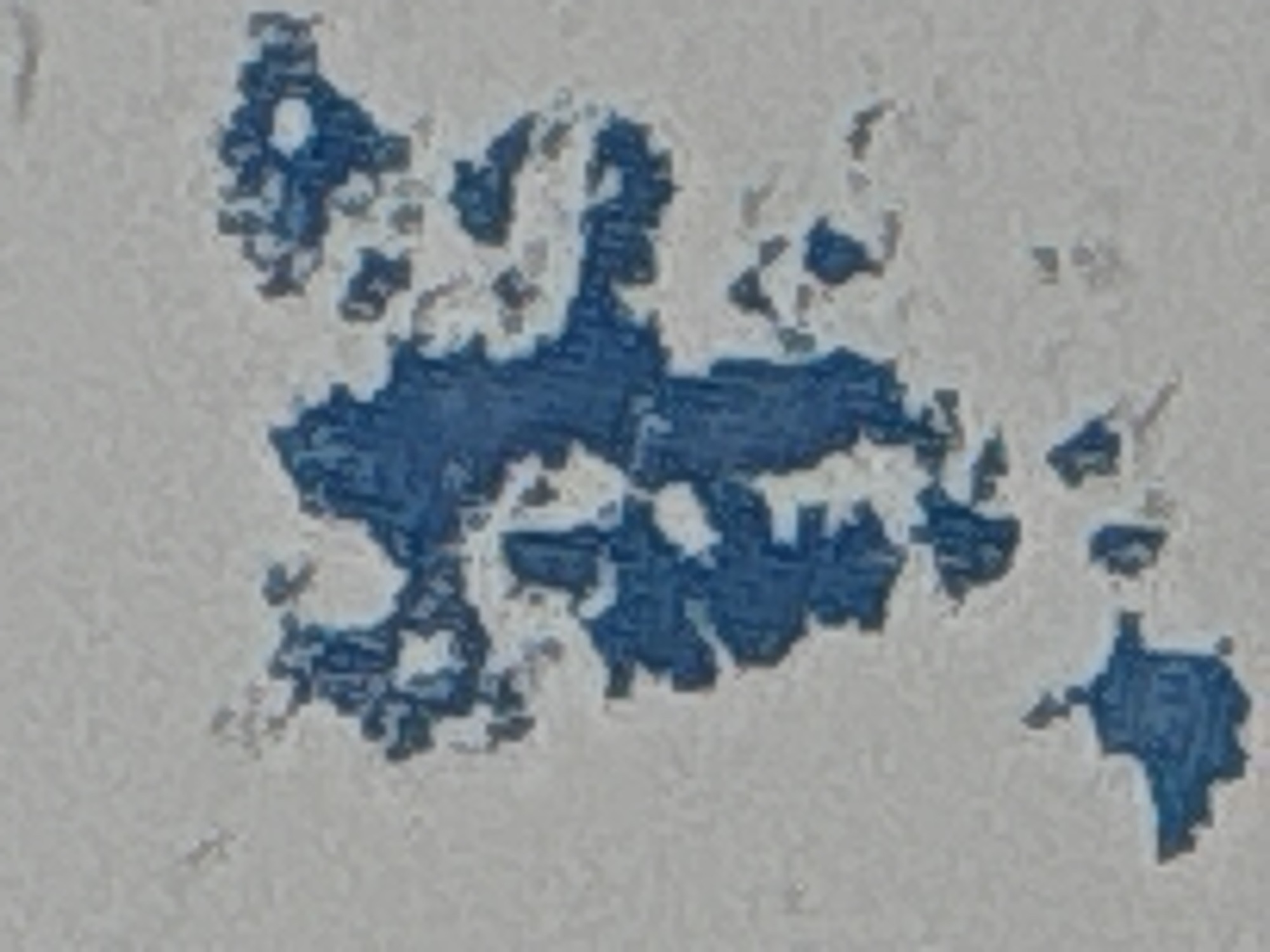 The image displays a close-up, abstract view of blue paint on a textured, light gray surface. The blue paint appears to be peeling or chipped, revealing the gray surface underneath in irregular patterns. The texture of the gray surface is granular, resembling concrete or a rough wall. The distribution of the blue paint is scattered and irregular, with larger patches and smaller flecks. The lighting is even, casting no distinct shadows, suggesting an indoor or overcast environment. The image lacks any discernible context of a city or specific location.